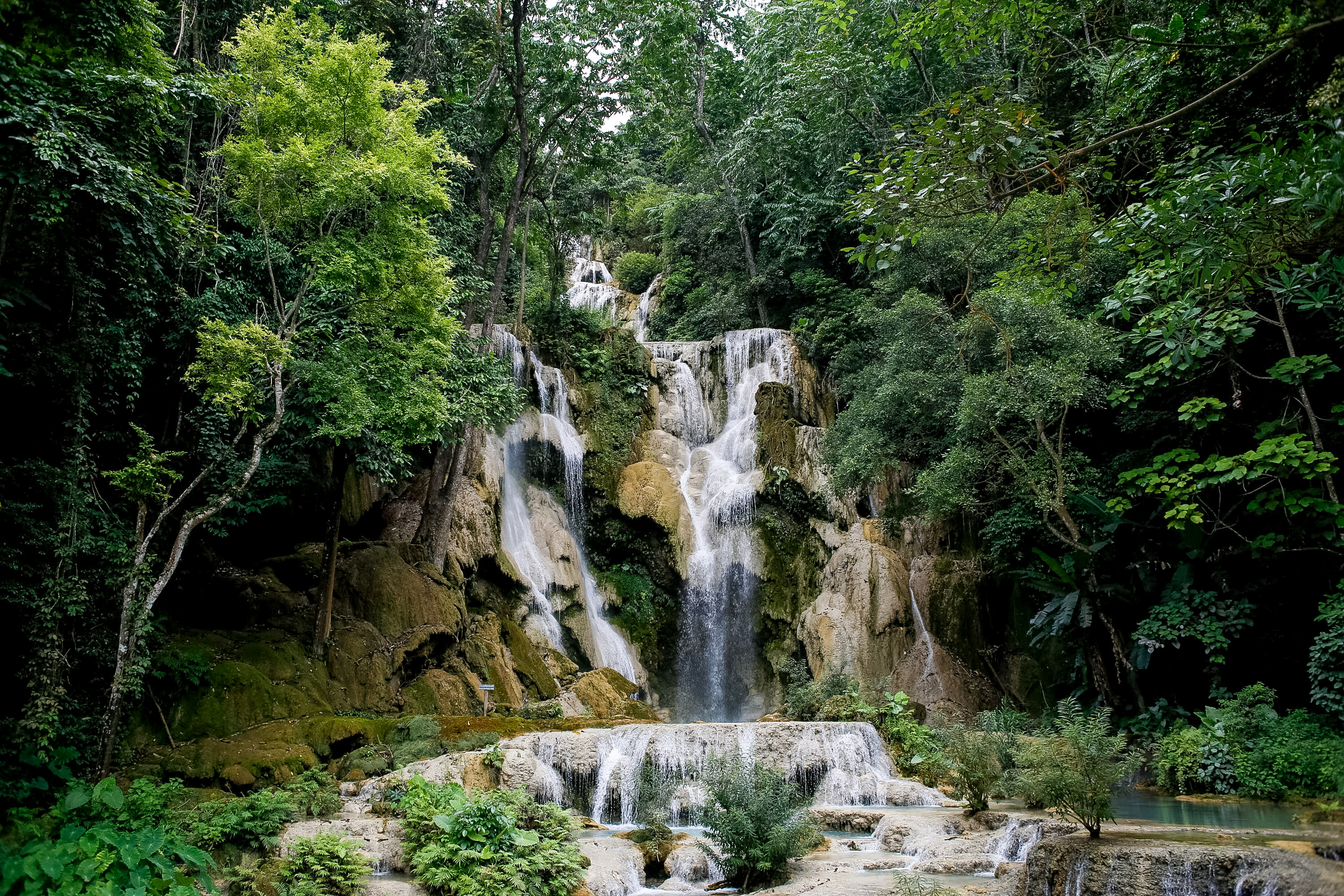 A wide, multi-tiered waterfall drops through limestone terraces into turquoise pools shaded by forest.