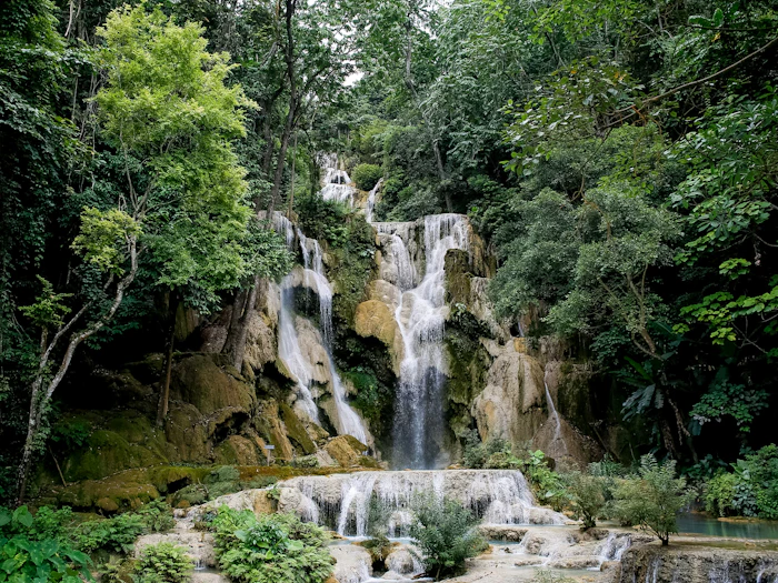 A wide, multi-tiered waterfall drops through limestone terraces into turquoise pools shaded by forest.