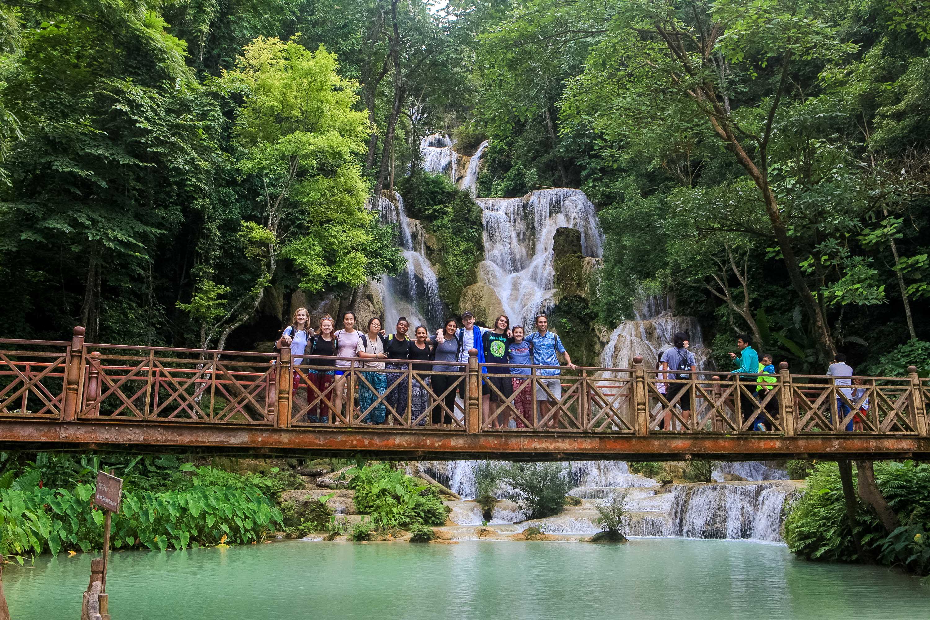 A group of students poses on a wooden footbridge with a tiered waterfall behind them.