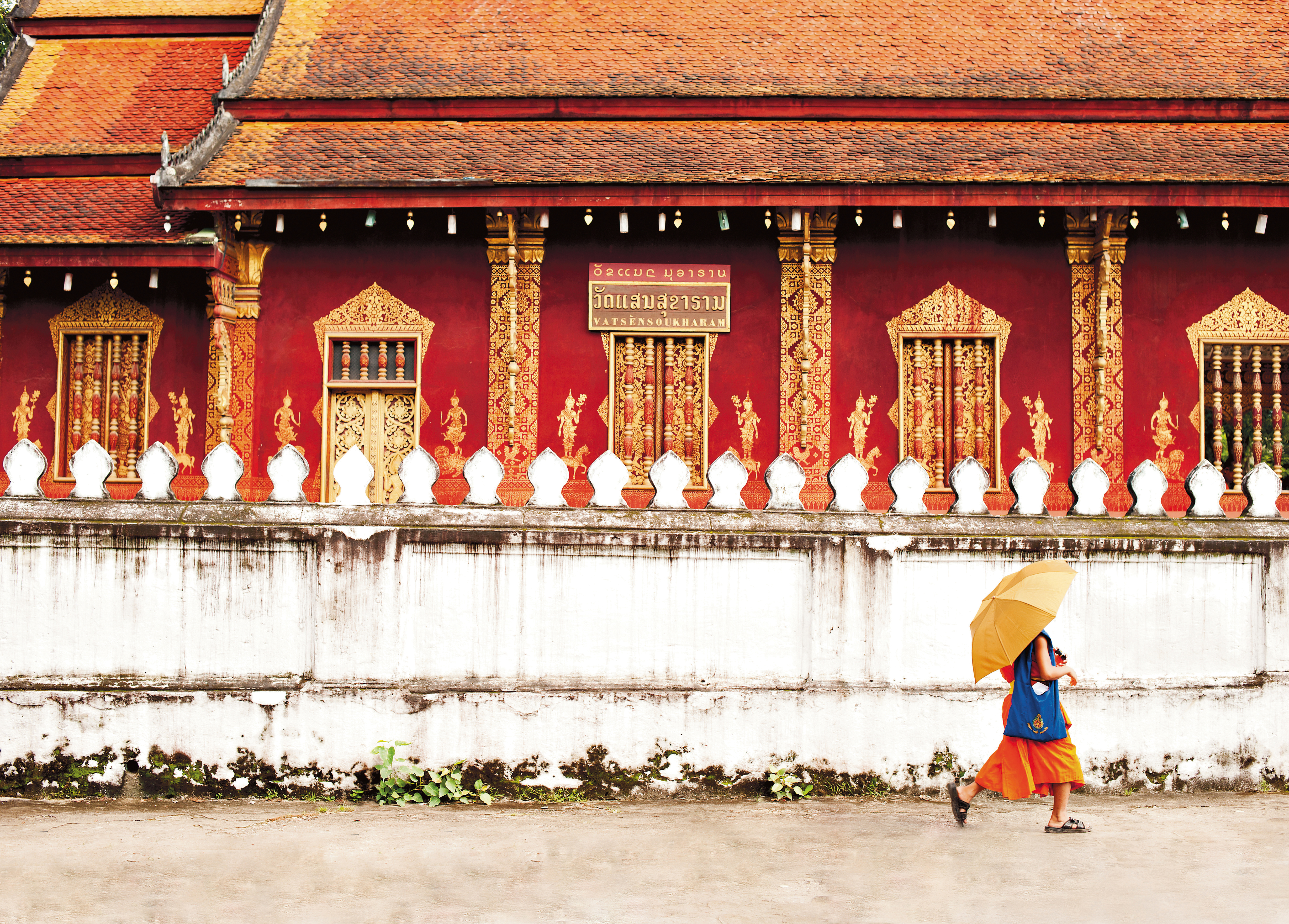 A novice monk in an orange robe stands before a small shrine platform in a wooded temple garden.