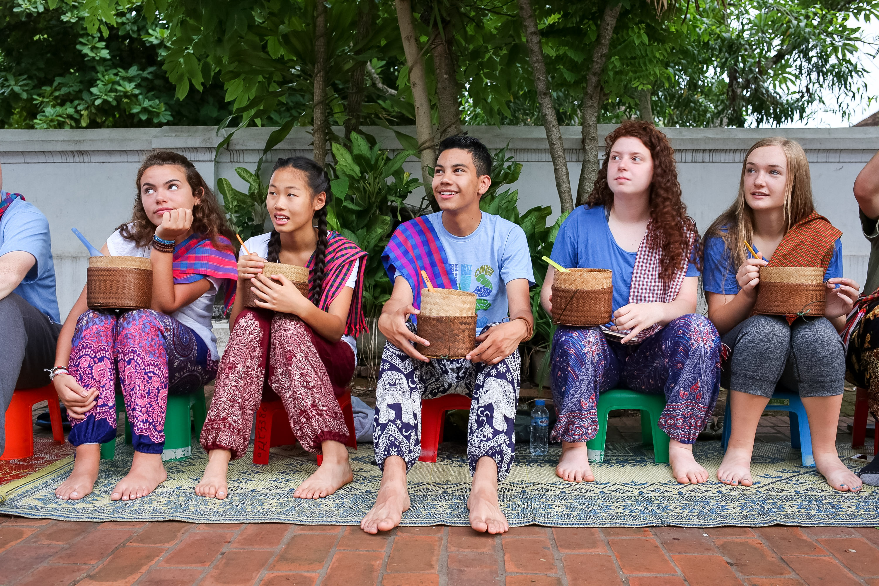 Students sit on stools holding woven baskets under shaded trees