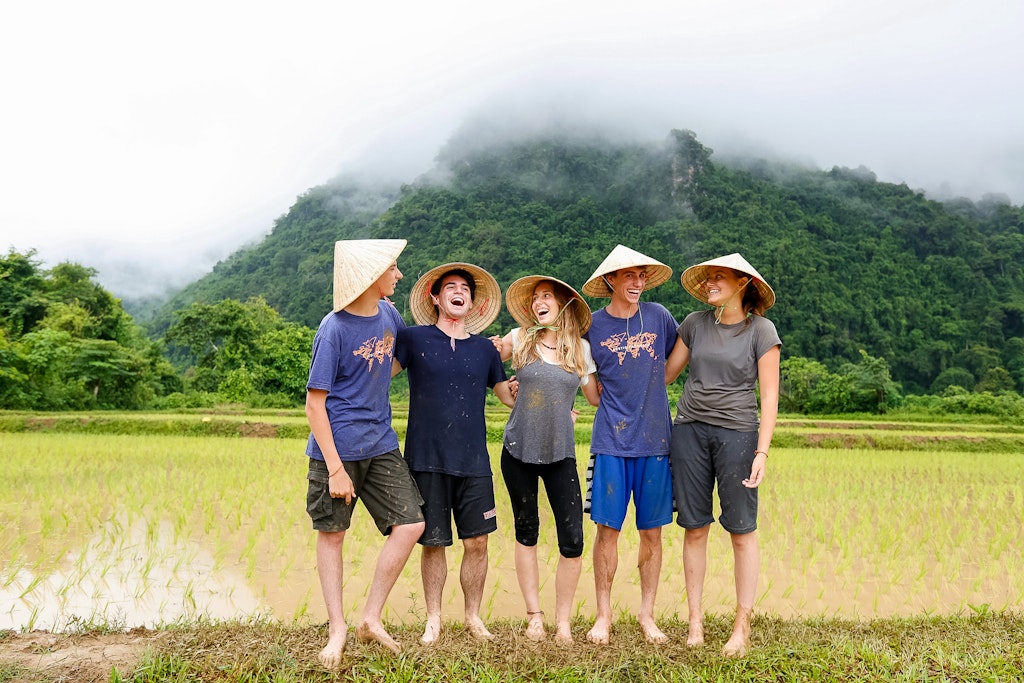 Four students stand in front of a field, wearing traditional hats from Laos.
