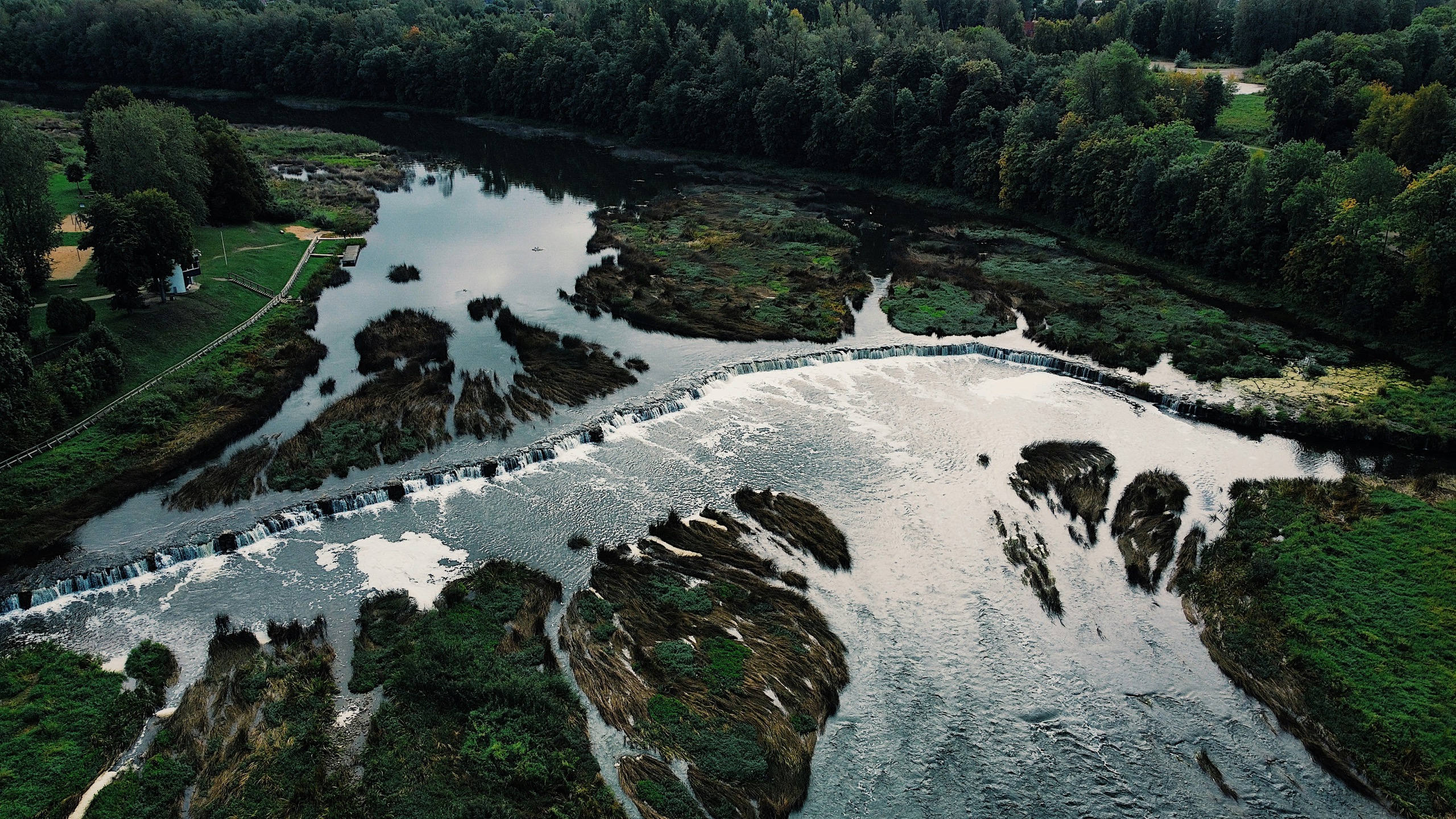 A wide, low waterfall spreads across a river between wooded banks, seen from above.