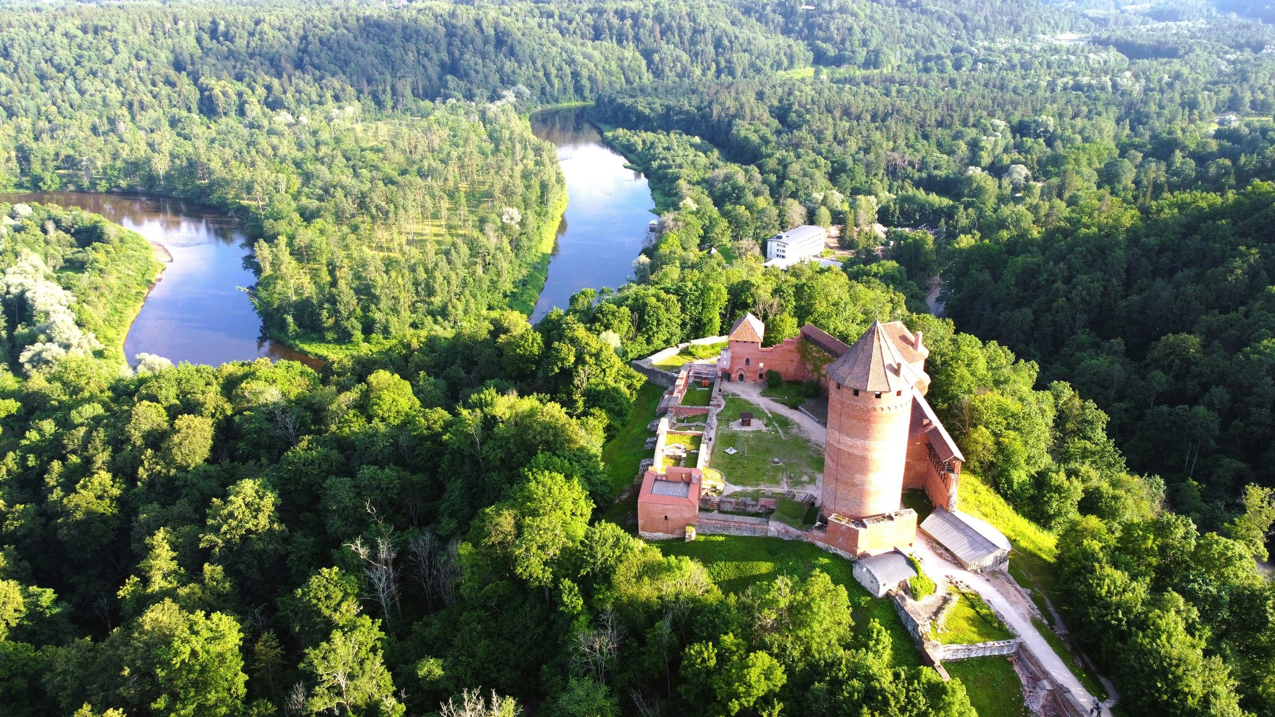 A red-brick castle with a round tower stands on a wooded hill above a bend in a river, seen from above.