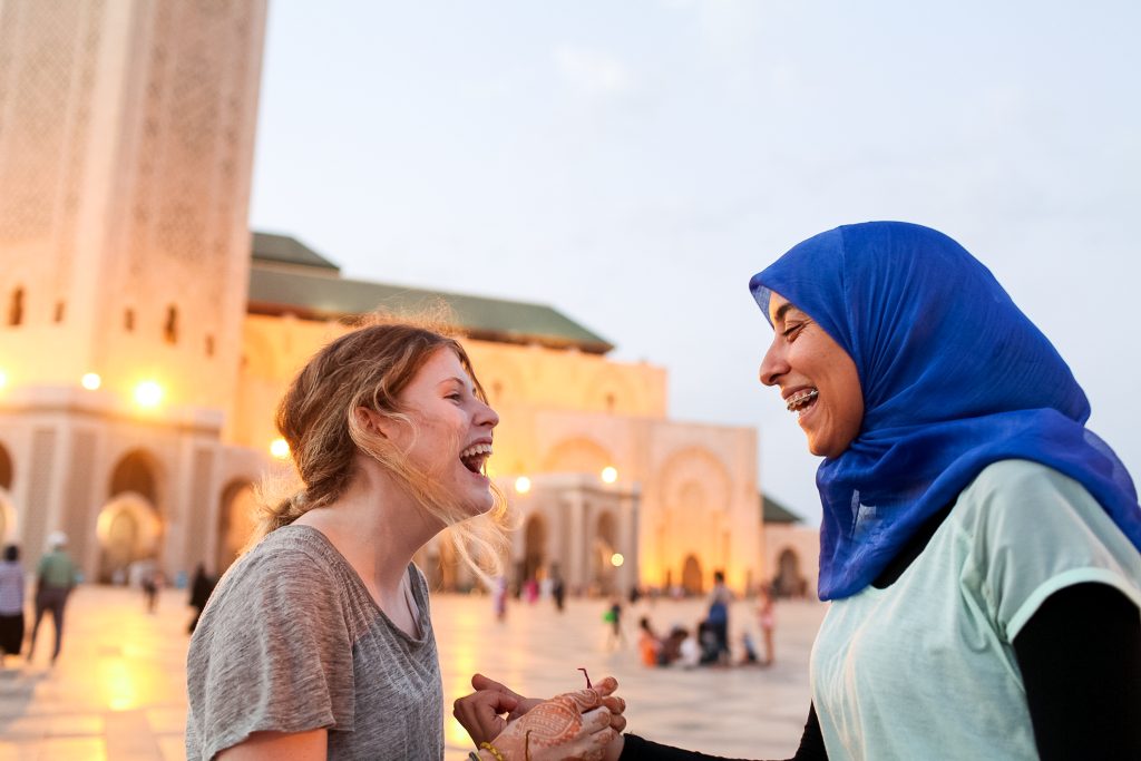 A student traveler and a local woman laughing while talking together in front of a mosque in Morocco.