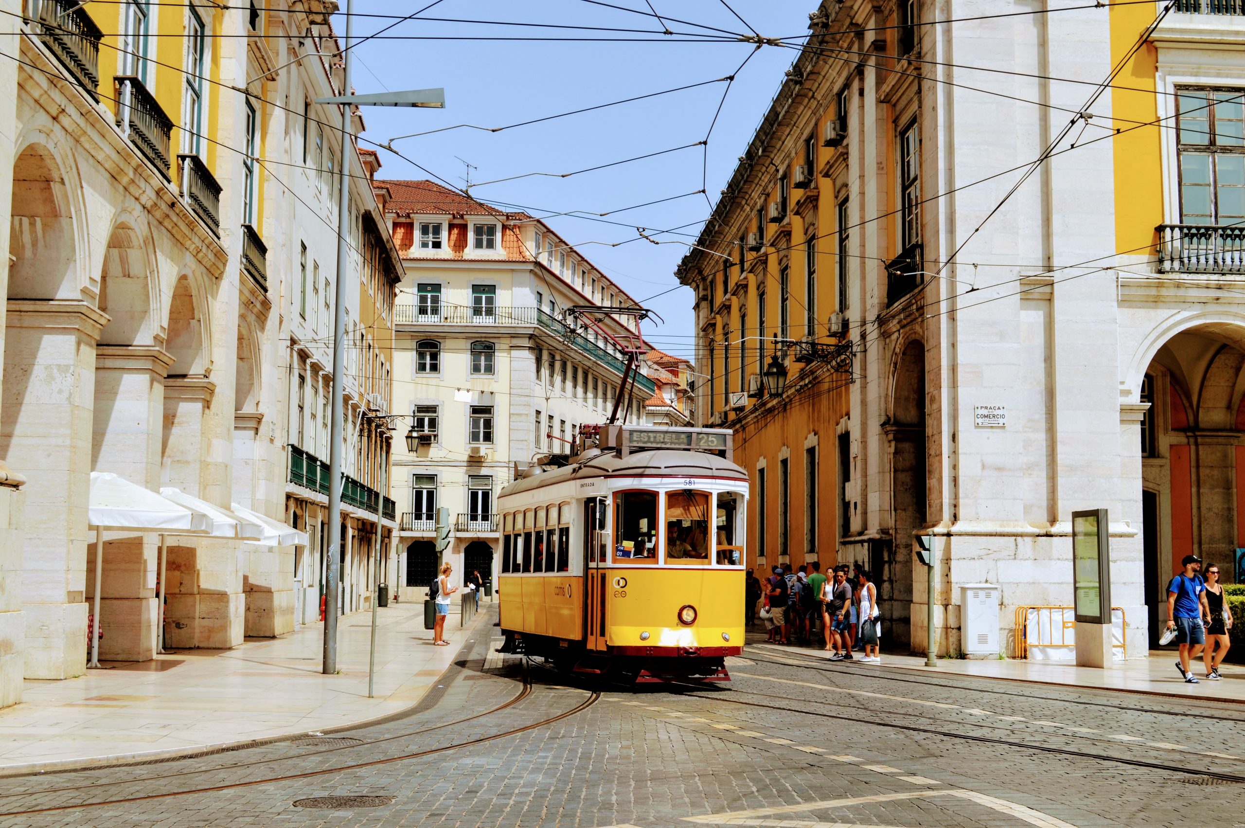 A yellow tram waits on cobbled tracks in a wide arcaded square, overhead wires crossing above it and pale stone buildings with arches on both sides.