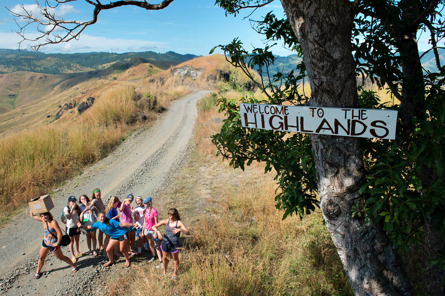 Teen travelers pose on a gravel road beside a "Welcome to the Highlands" sign overlooking rolling hills