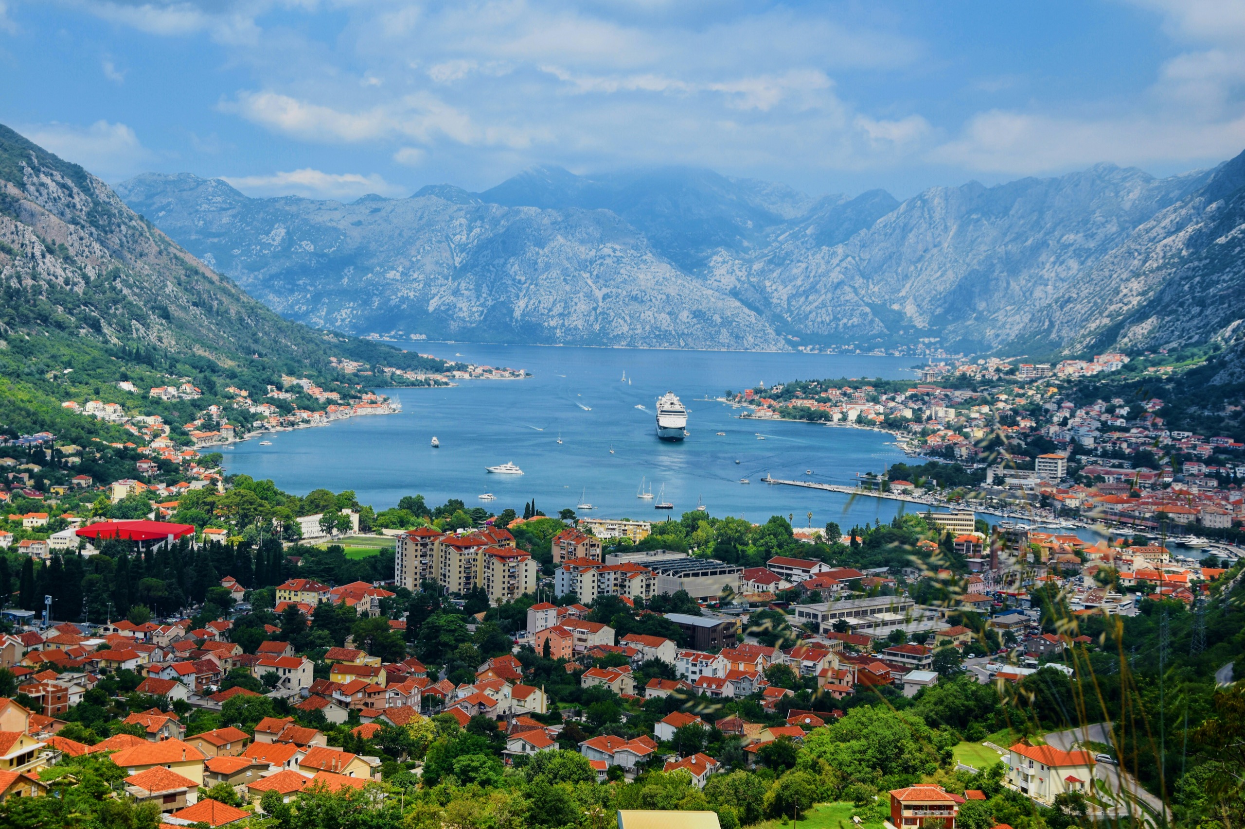 A view down the mountain-ringed Bay of Kotor, with a cruise ship at anchor and a marina along the shore.