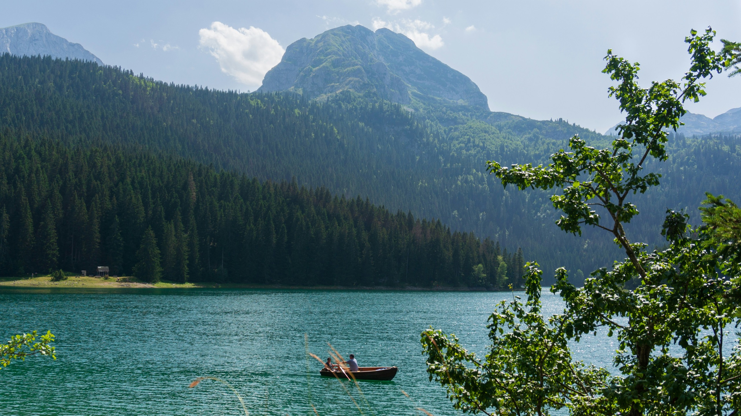 A single wooden rowboat with two people on a still green lake, backed by pine forest climbing to a bare grey peak.