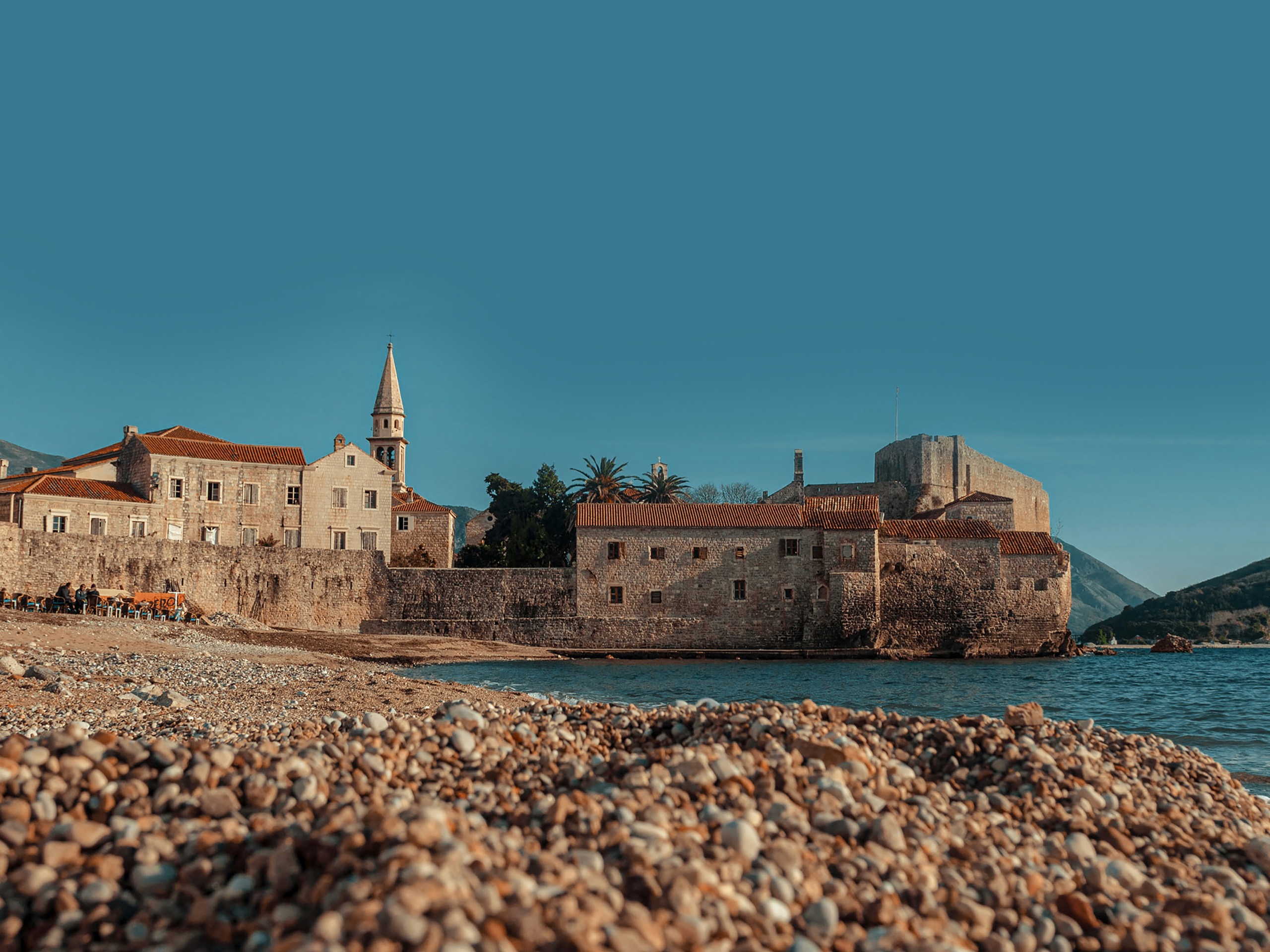 Budva's walled old town seen from the pebble beach, its bell tower and tiled roofs above the waterline.