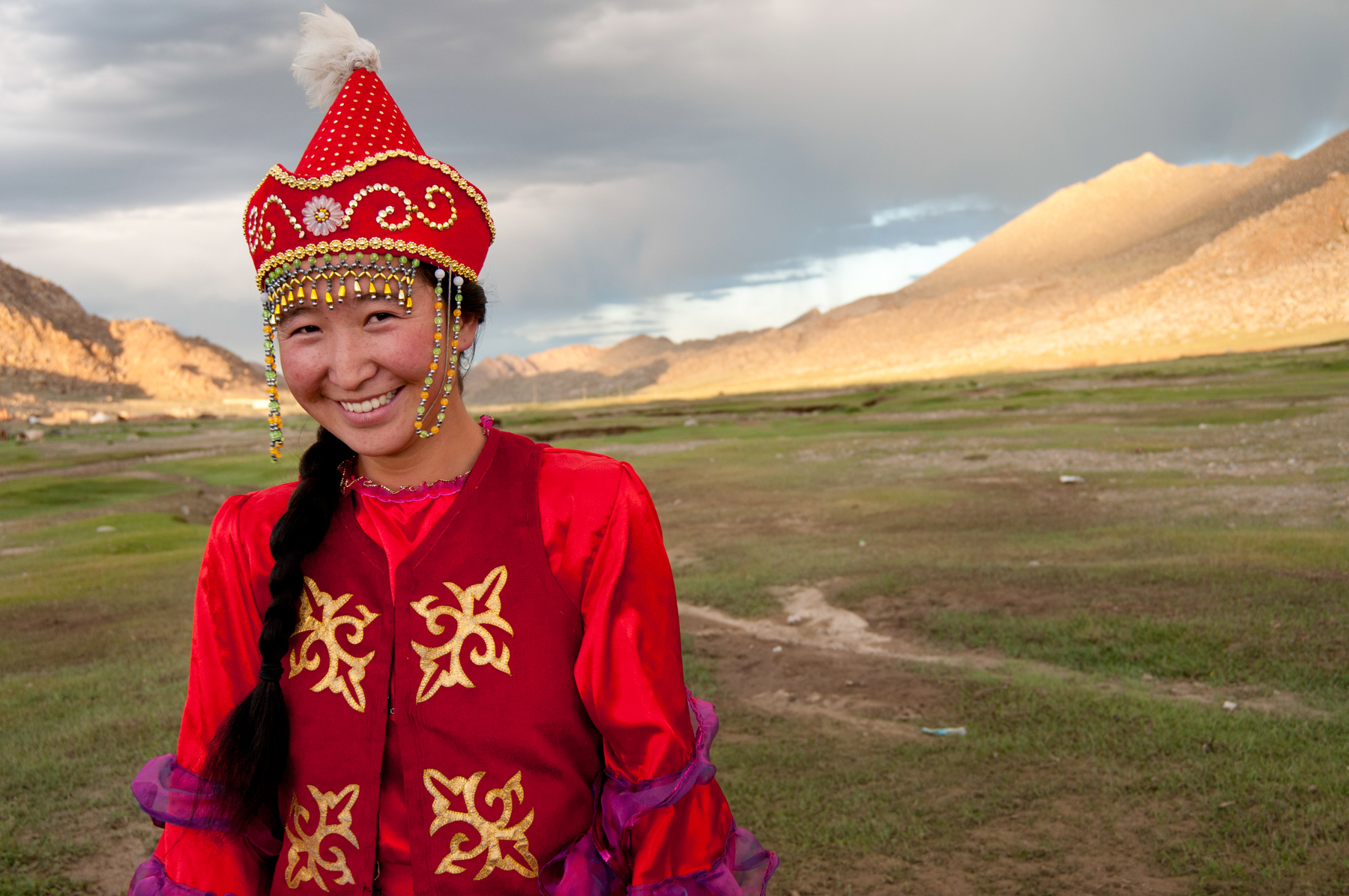 A woman in traditional Mongolian dress and pointed hat smiles in front of grassy hills in Mongolia