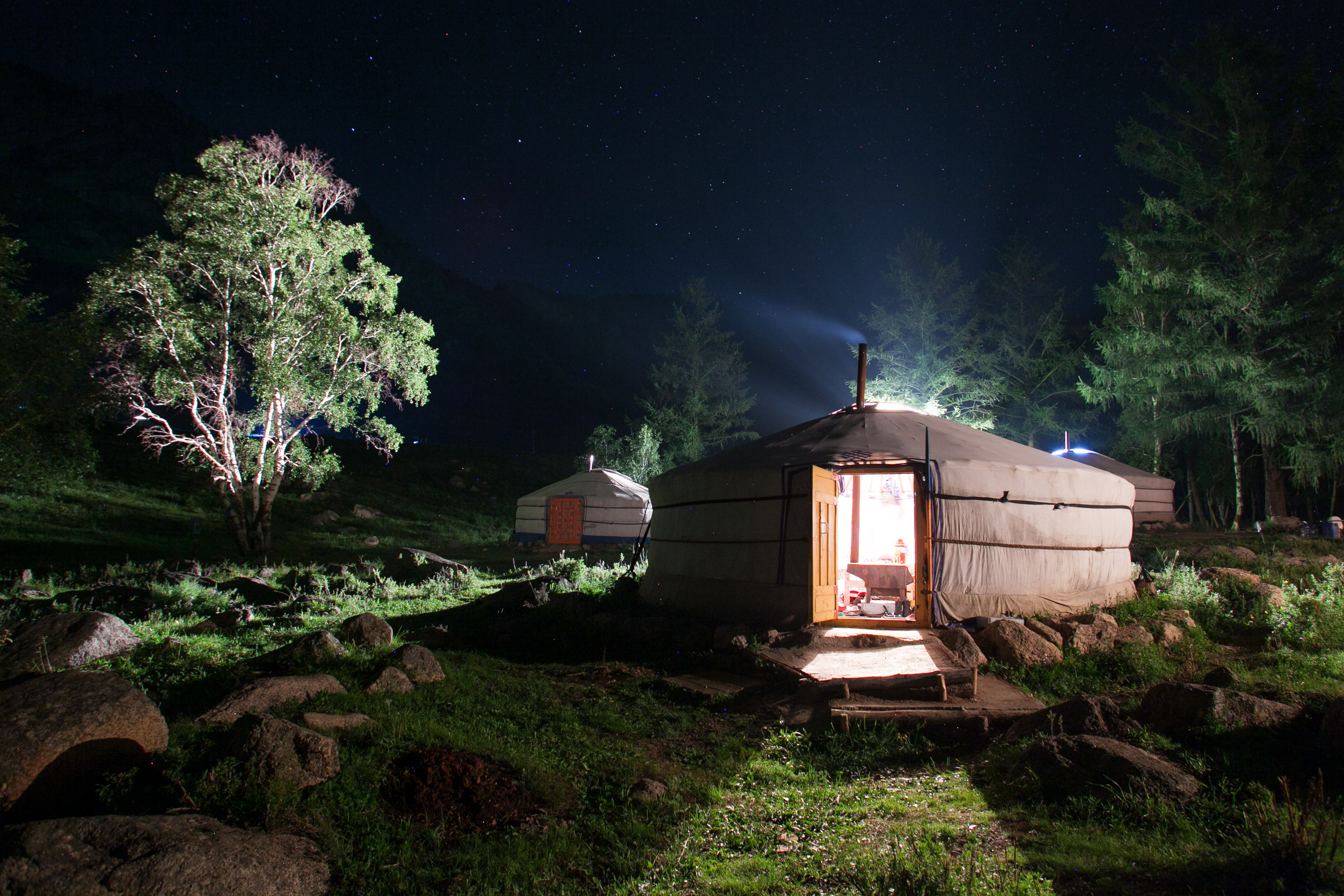 A lit ger glows against a starry night sky with silhouetted trees and rocky terrain