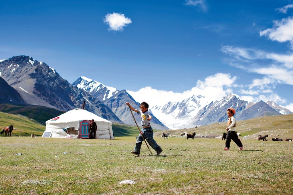 Two children play outside on a grassy plain in Mongolia, with a yurt and horses in the background. Snow-capped mountains rise majestically under a bright blue sky, scattered with a few clouds.