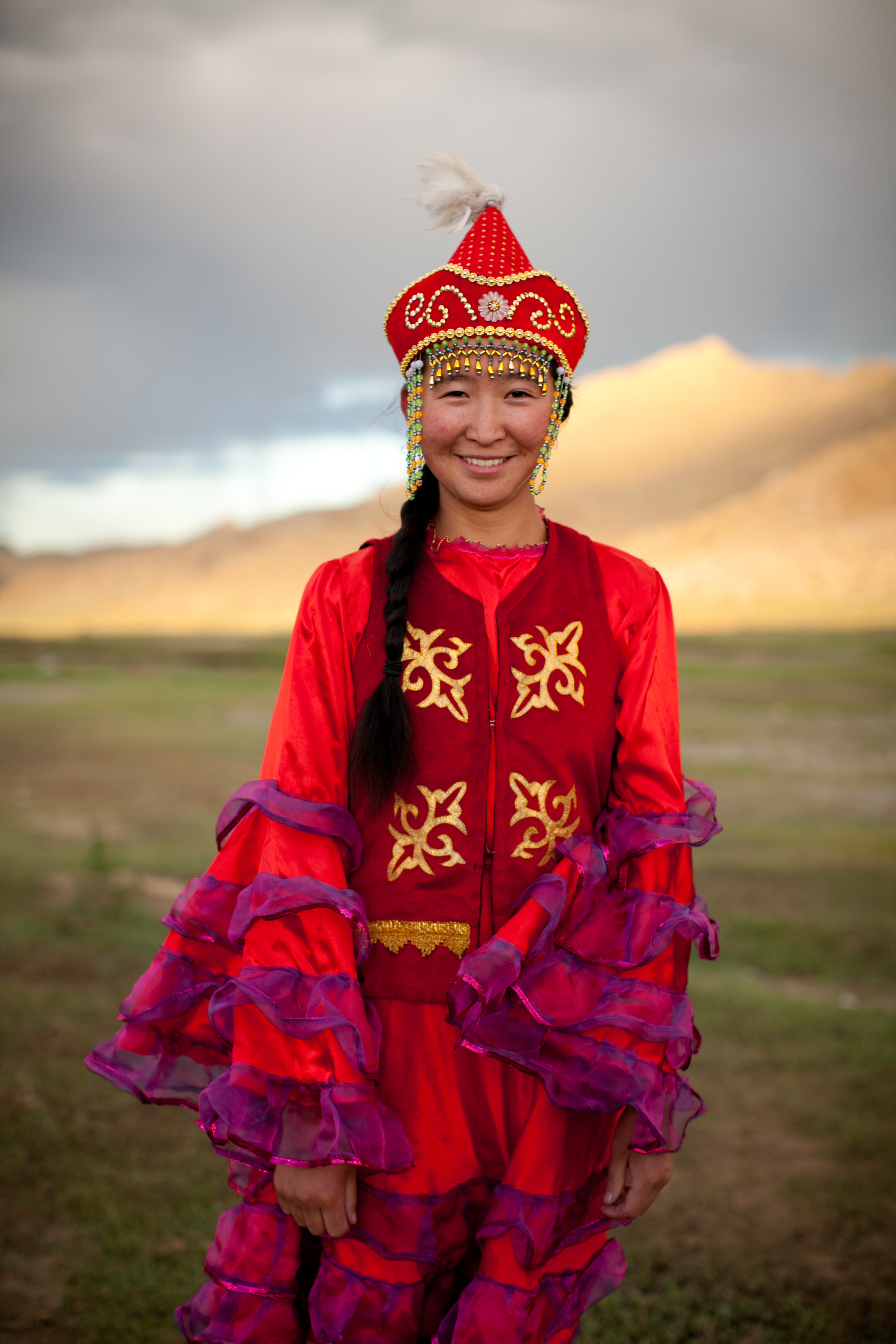 A woman poses in traditional Mongolian dress with a peaked hat against a mountain backdrop