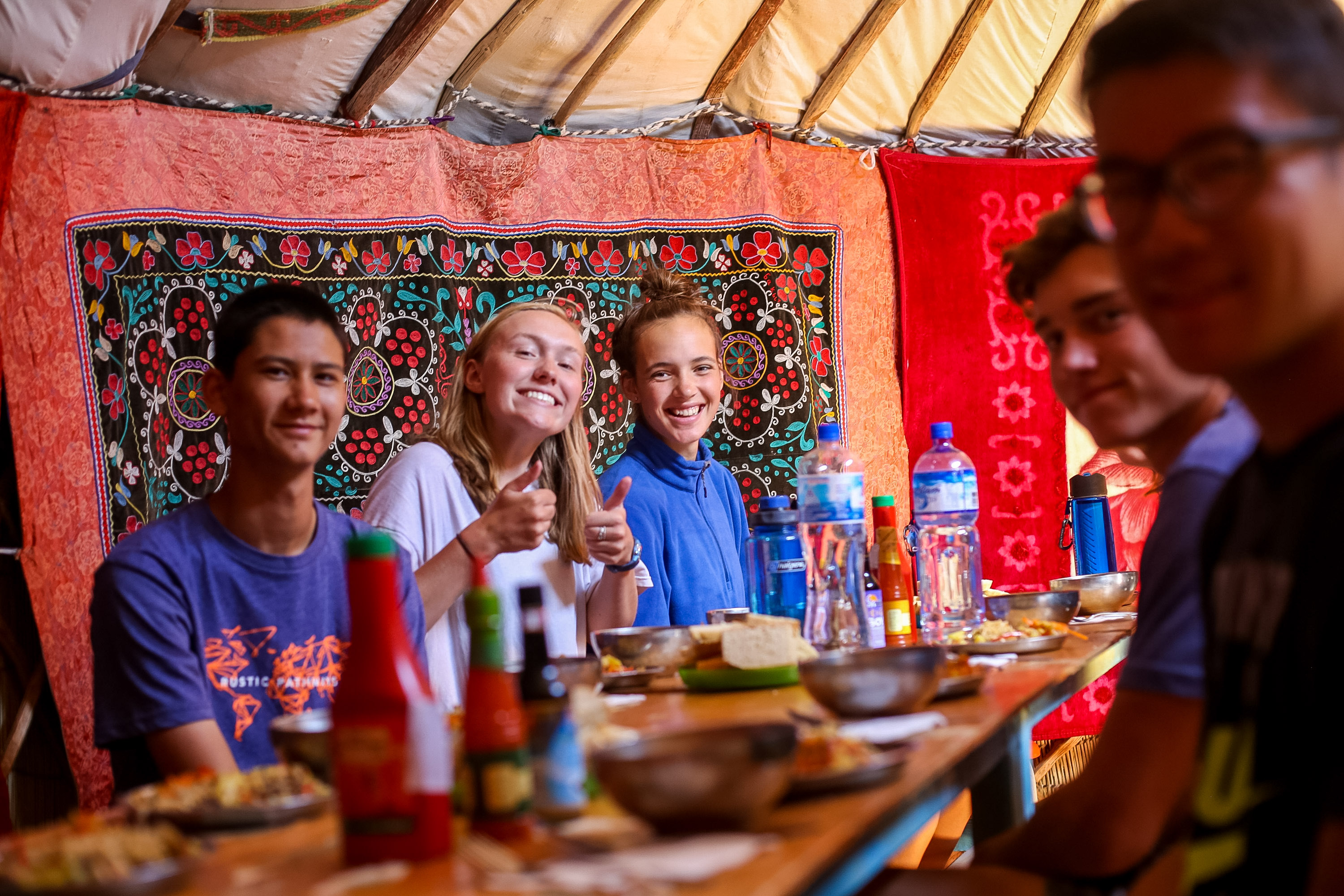Students sit along a long table inside a ger, in front of a bright patterned wall hanging, with bowls and bottles set out for a meal.