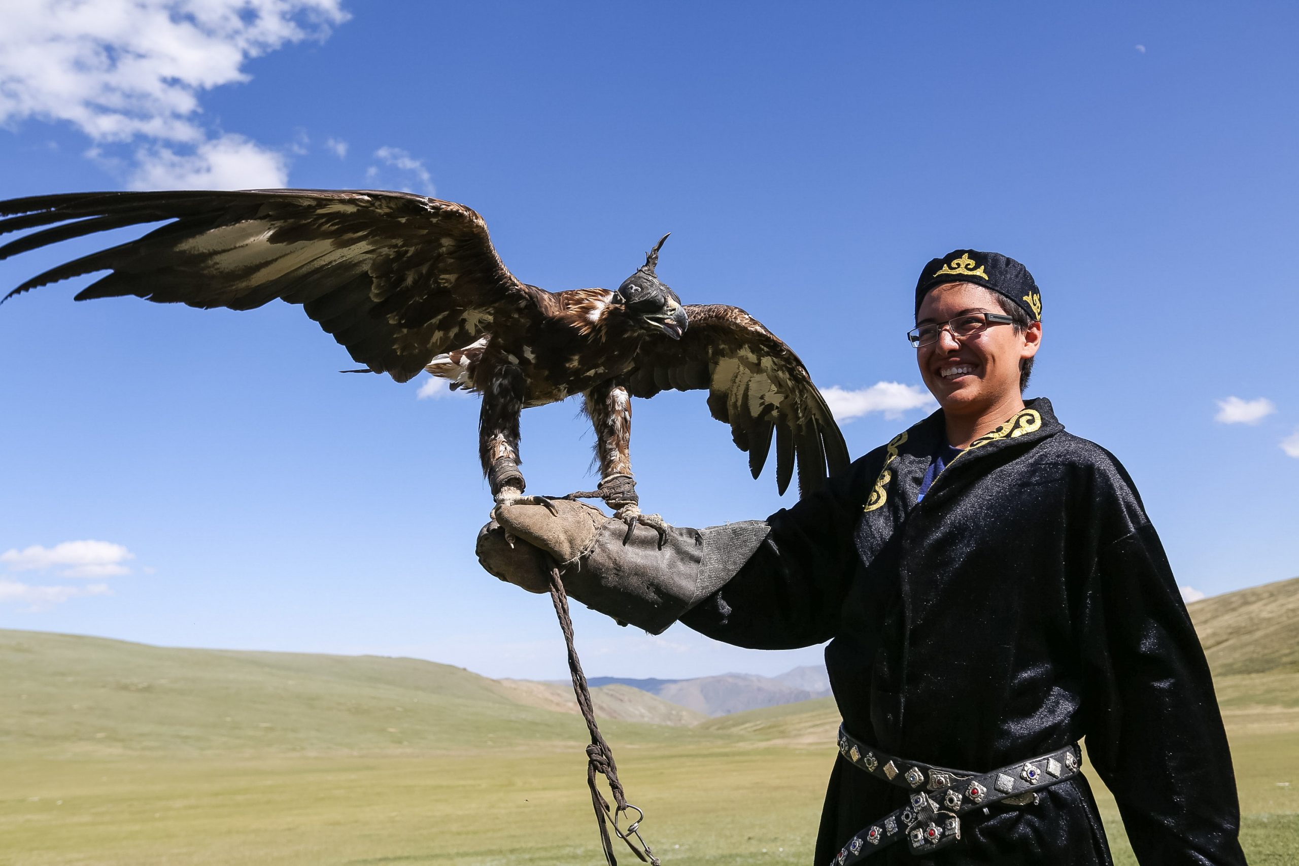 A Kazakh eagle hunter in a black embroidered coat stands on open steppe holding a golden eagle on his gloved arm, the bird's wings spread wide, with bare hills rising behind him.