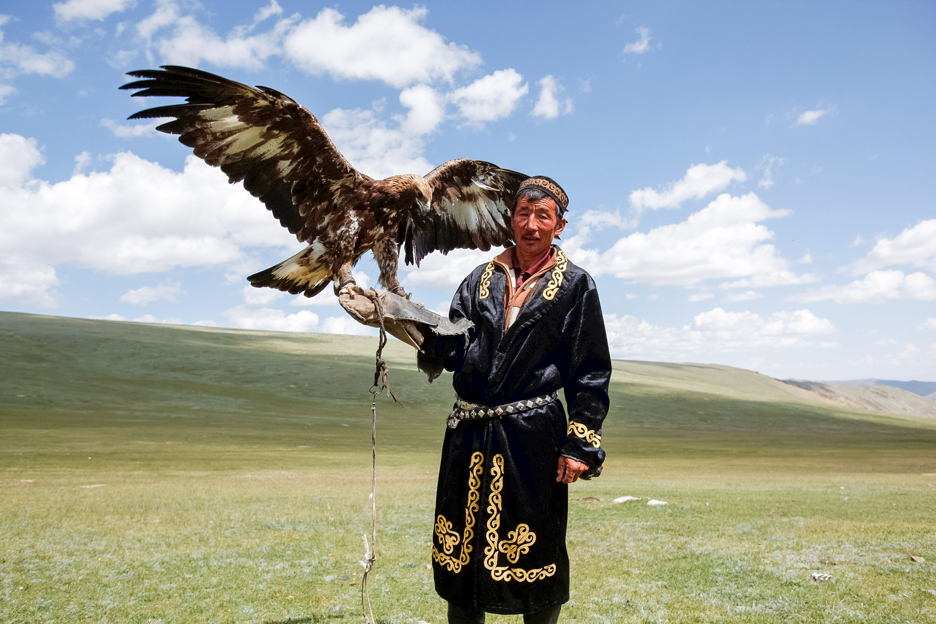 A man in traditional Mongolian dress holds a golden eagle on his arm in an open grassland as students look on