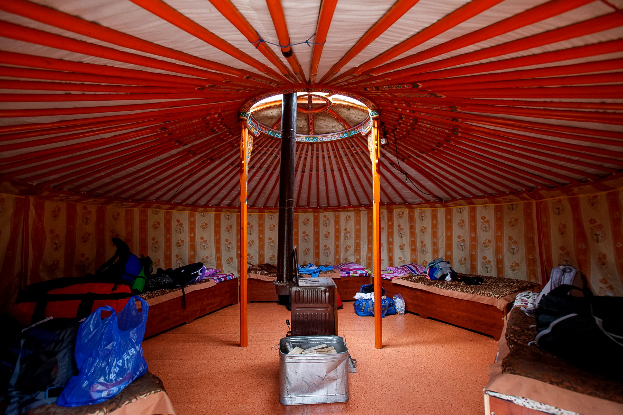 The inside of a ger: painted orange roof poles running to a central ring, a low stove in the middle, and simple beds with folded bedding set around the wall.