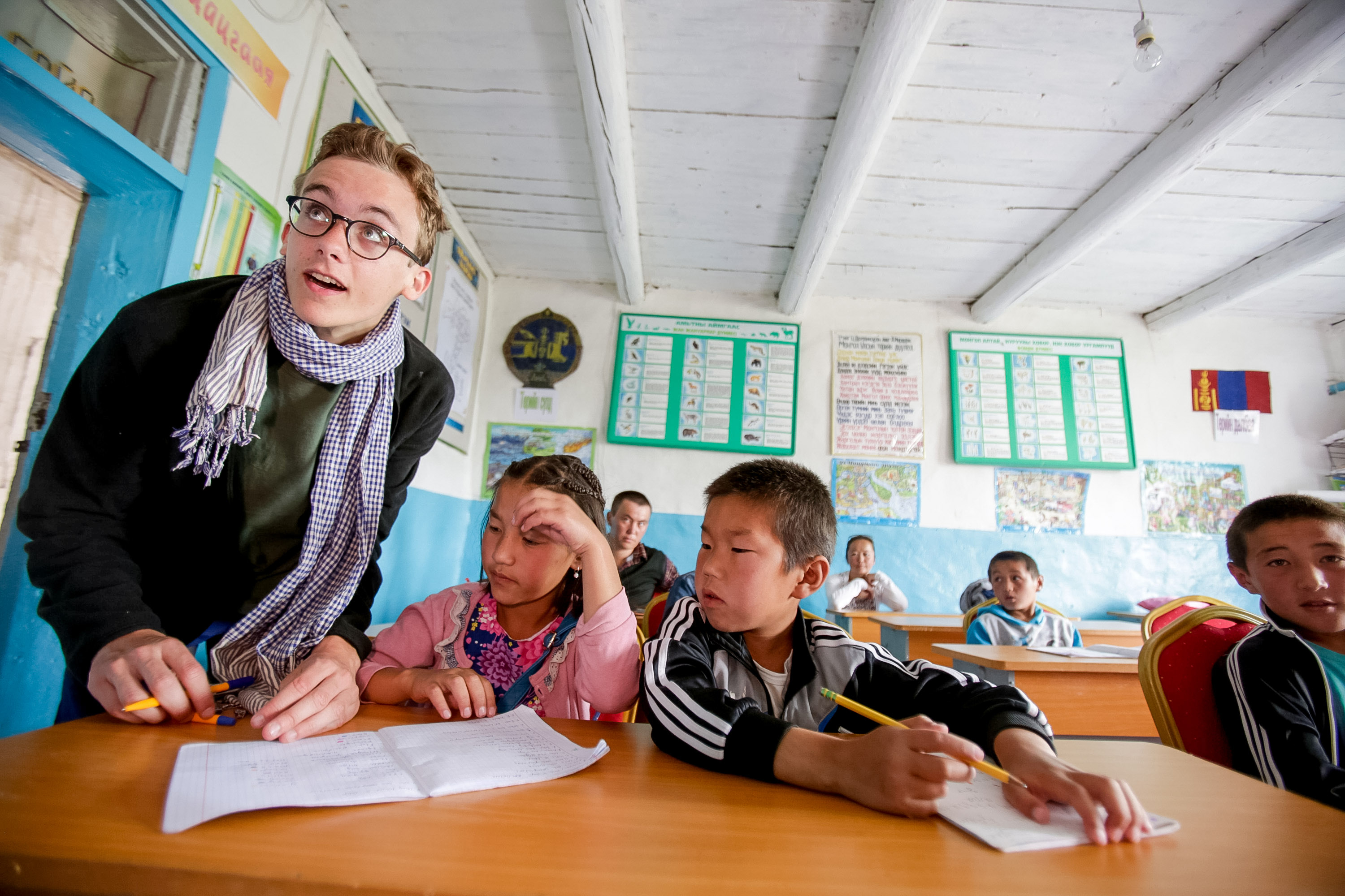 A leader leans over a classroom desk helping local students with their schoolwork
