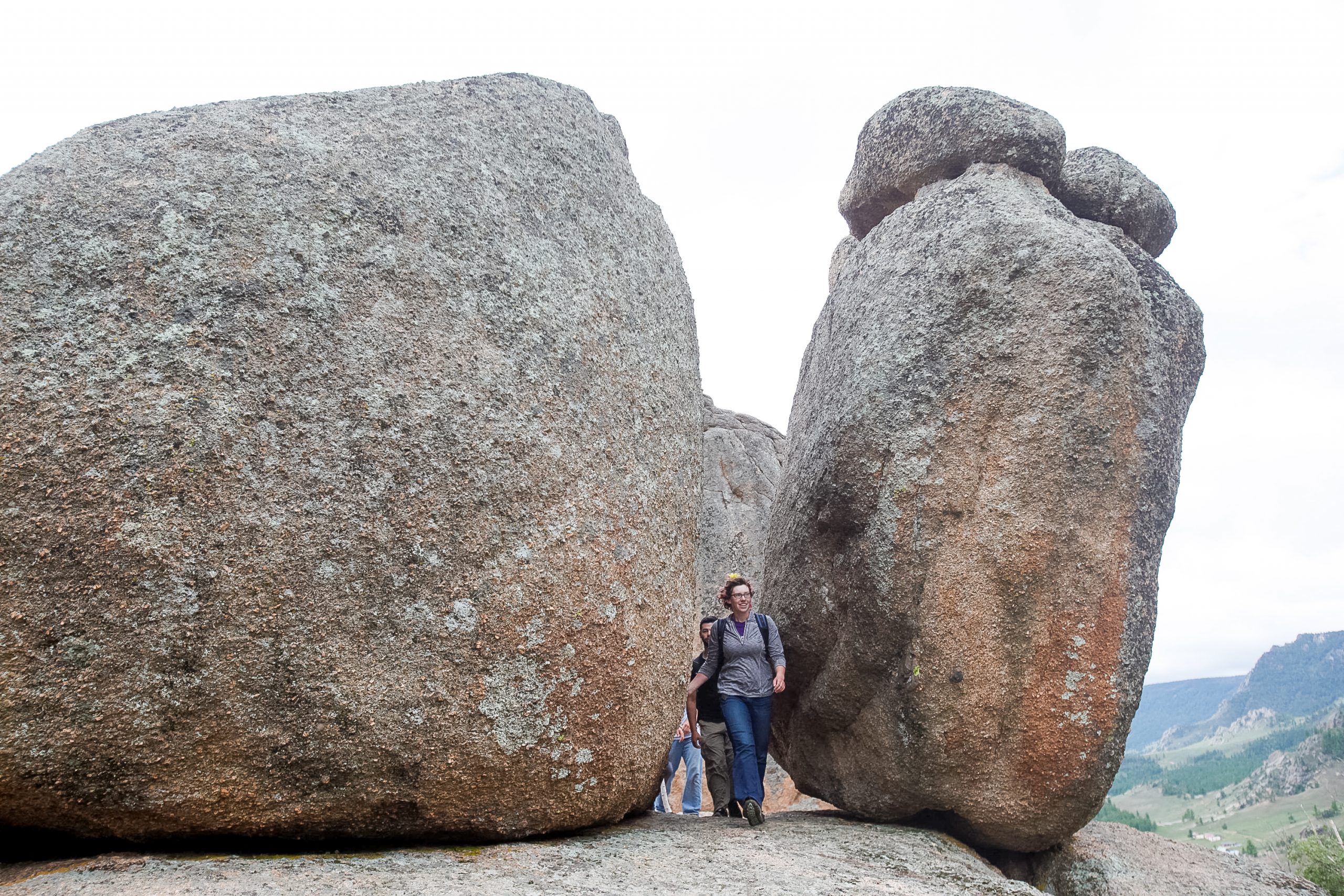 Two rounded granite boulders the size of small buildings stand a few steps apart on a grassy slope, with several people walking through the narrow gap at their base and open hills running away behind them.