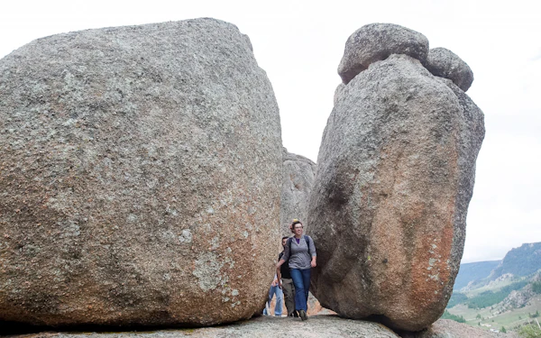 Two rounded granite boulders the size of small buildings stand a few steps apart on a grassy slope, with several people walking through the narrow gap at their base and open hills running away behind them.
