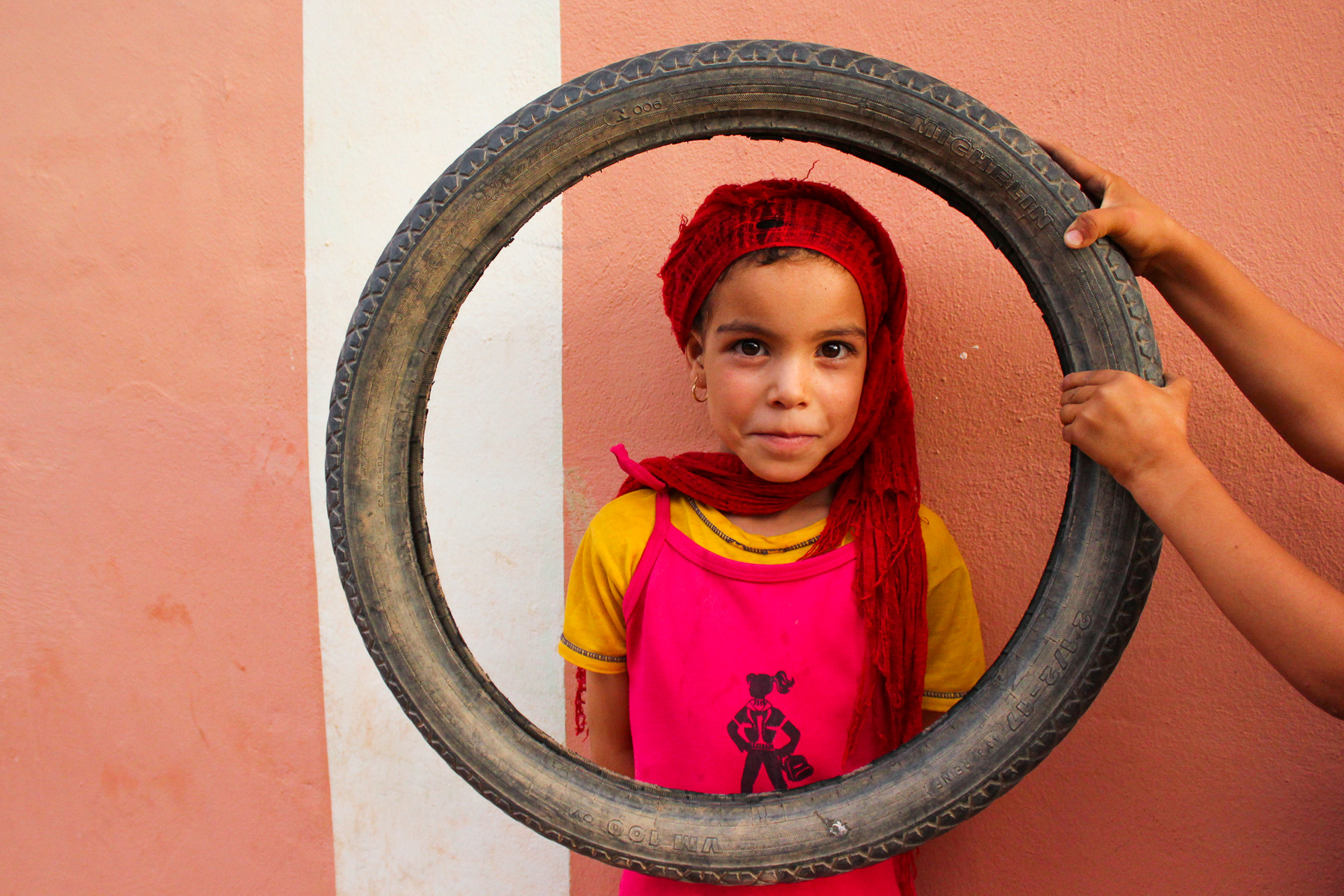On a school trip, a child in a red headscarf peers through a tire held by another person's hands