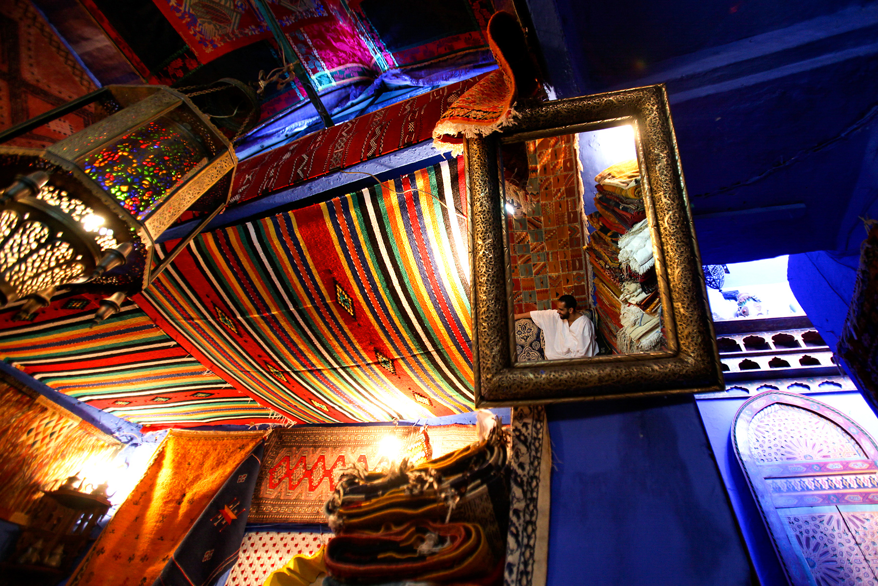 A mirror reflects a shopkeeper amid colorful woven rugs and lanterns in a Moroccan souk stall