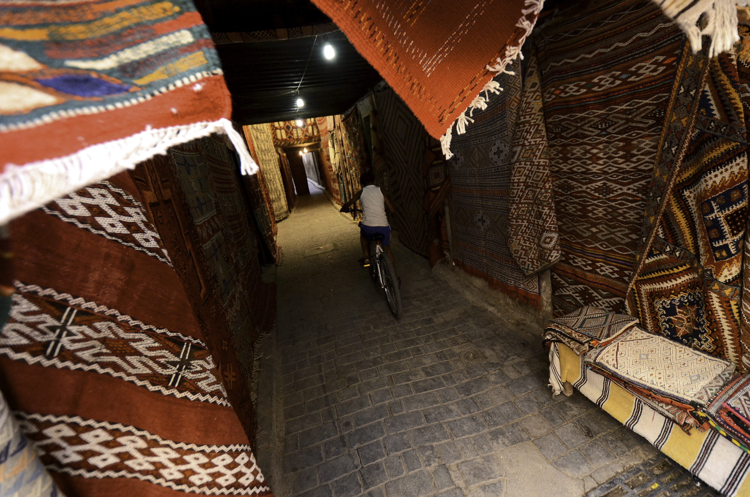 A cyclist rides through a narrow souq alley lined with hanging woven rugs