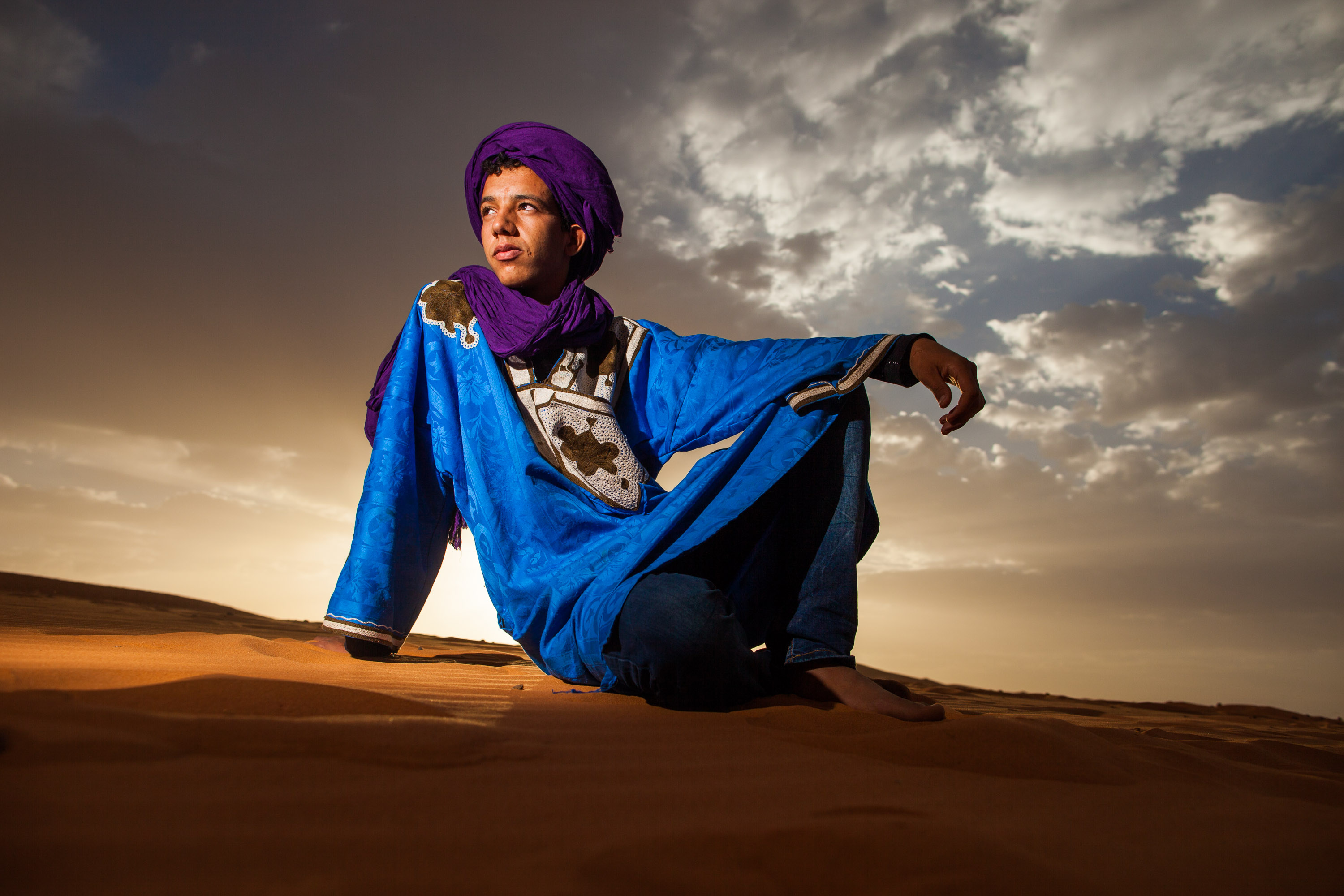 Students in traditional desert robes and turbans sit atop a Sahara sand dune at dusk