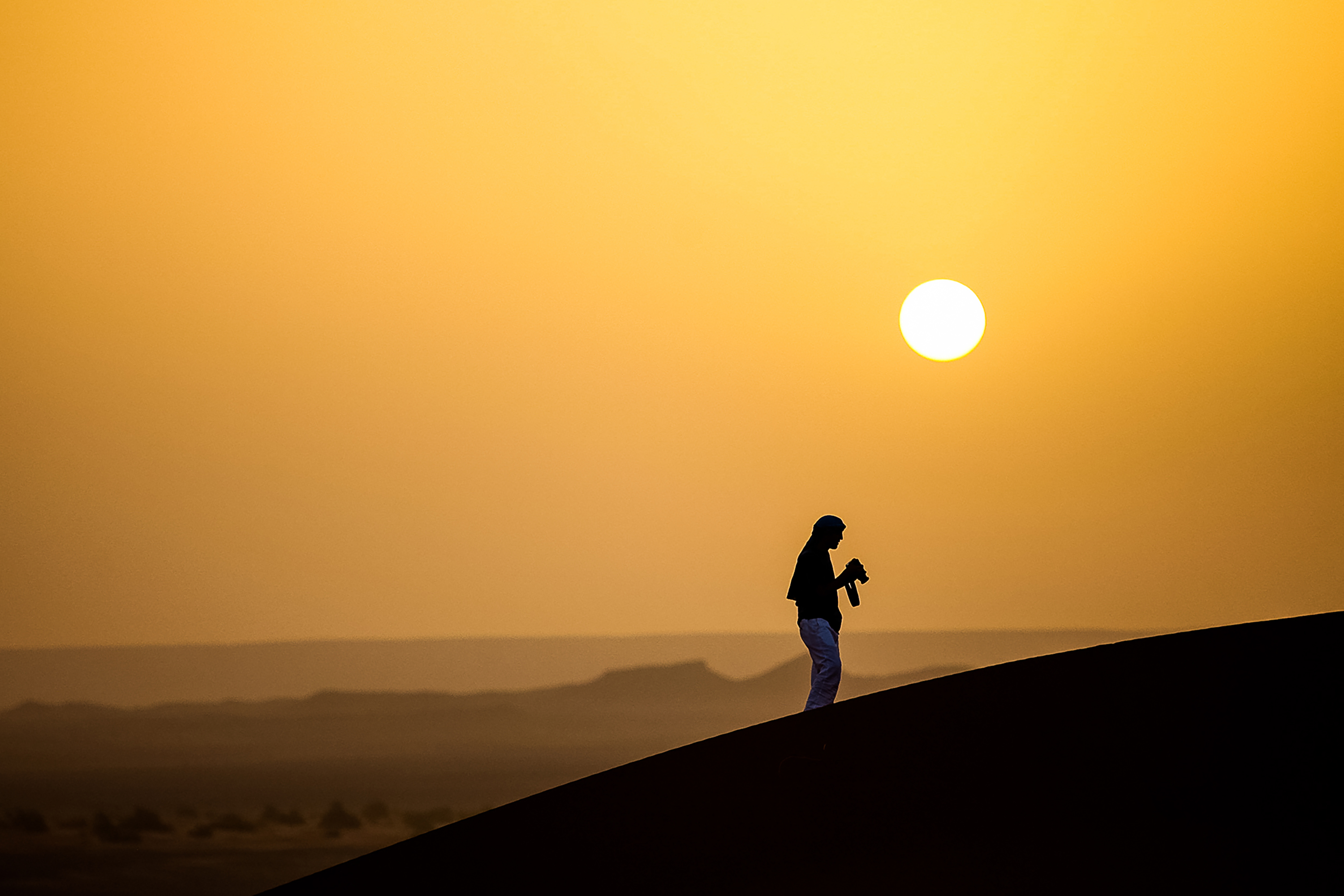 Silhouette of a person holding a camera on a sand dune at sunset.