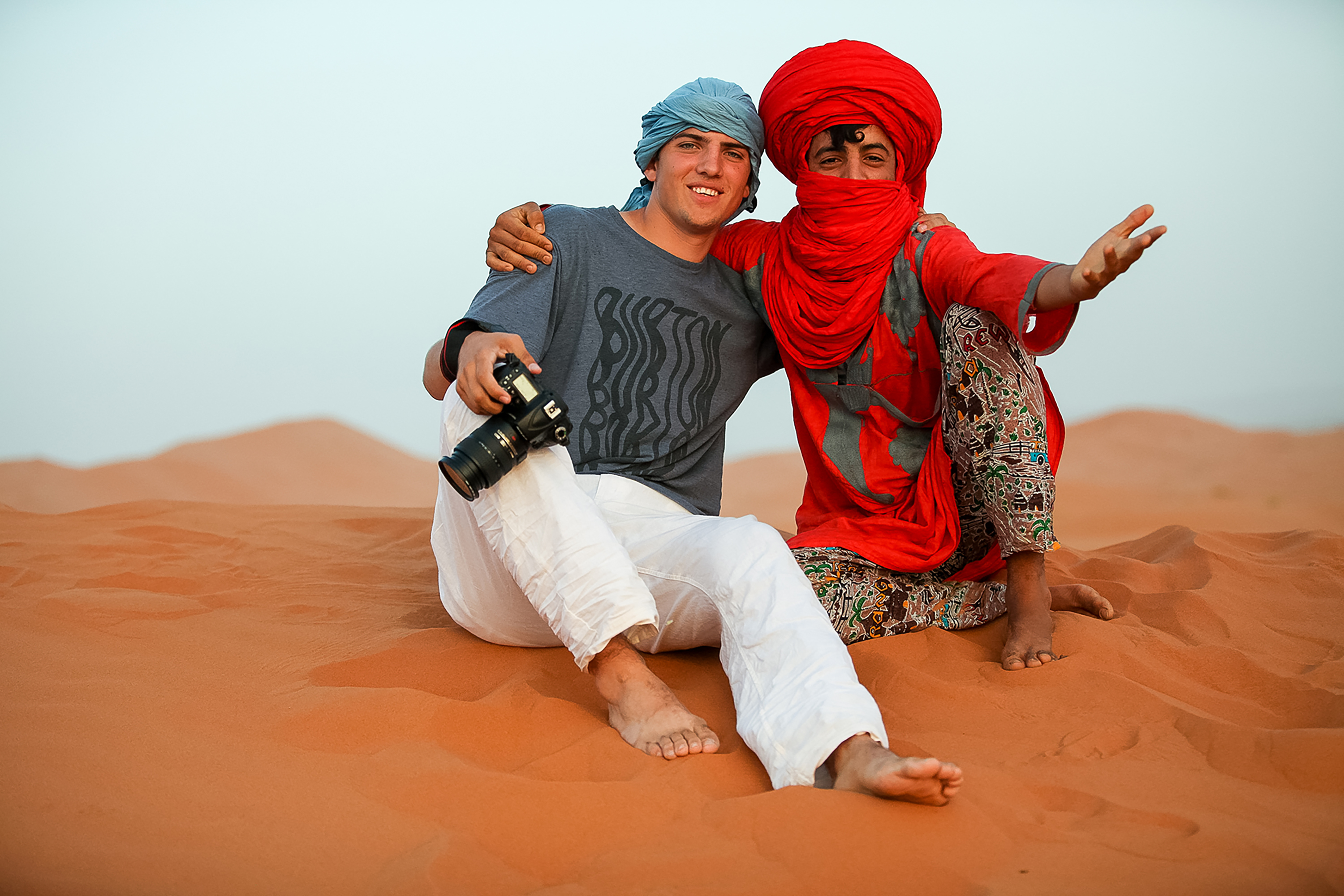A tourist and a local Moroccan man dressed in traditional clothing sit together on the sand dunes, smiling at the camera.