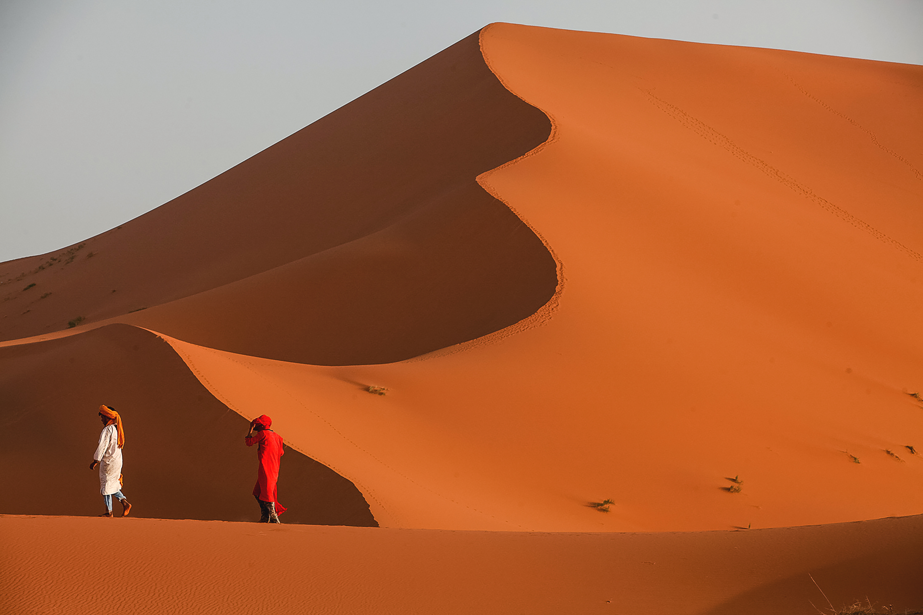 Teen travelers in flowing robes walk along the ridge of a towering desert sand dune