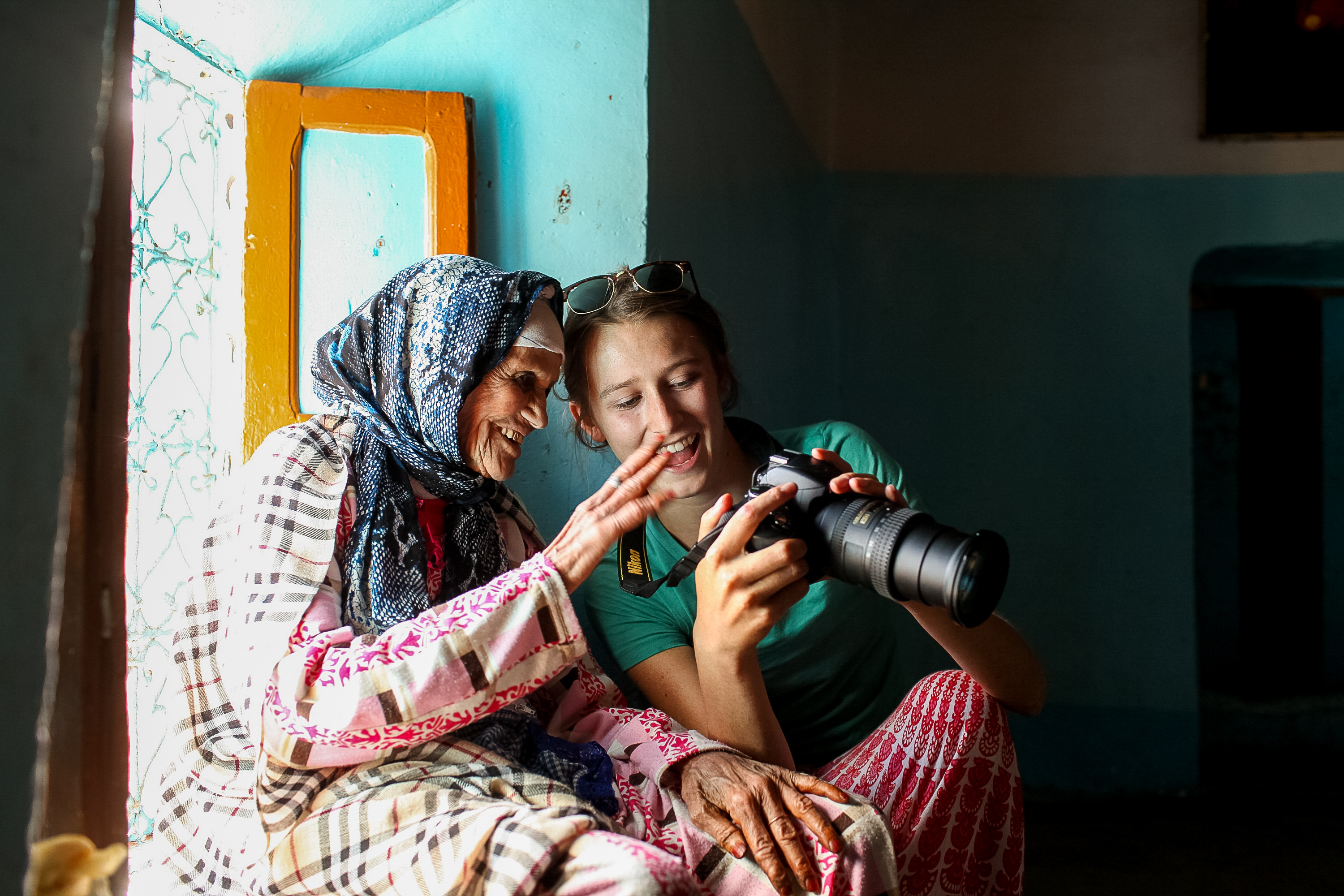 A student shows an older woman in a patterned headscarf the picture on the back of a camera, and both laugh as they sit together beside a turquoise wall.