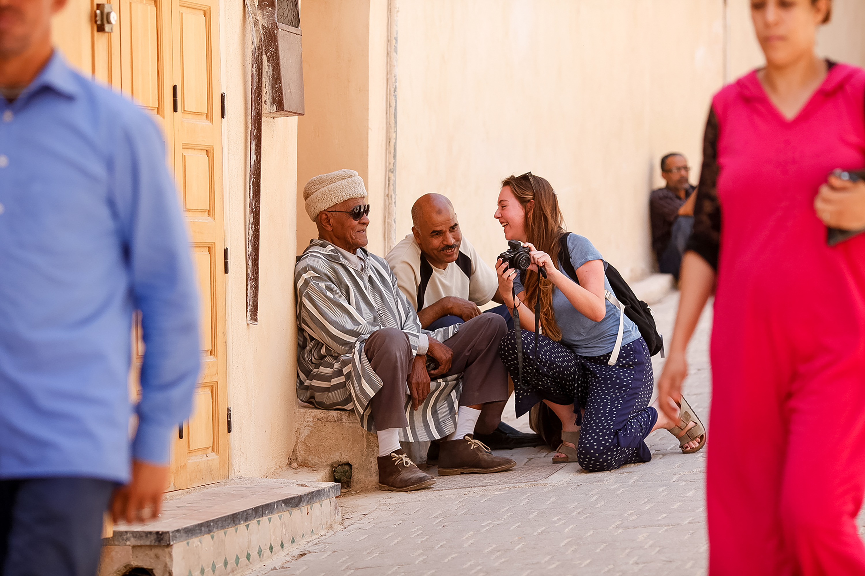 A student kneels to photograph local men sitting along a shaded medina wall