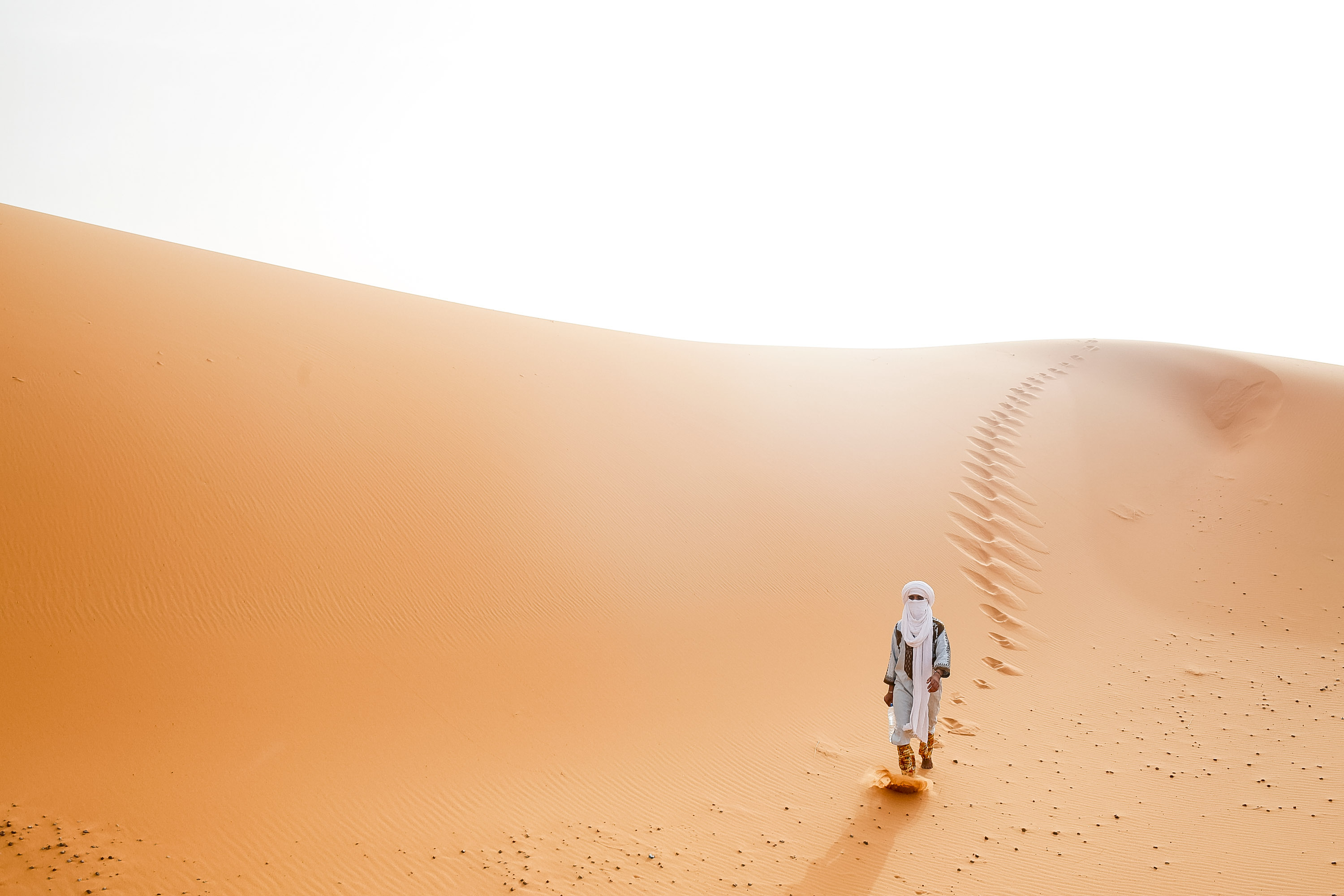 A student in traditional desert dress walks along a Sahara dune ridge