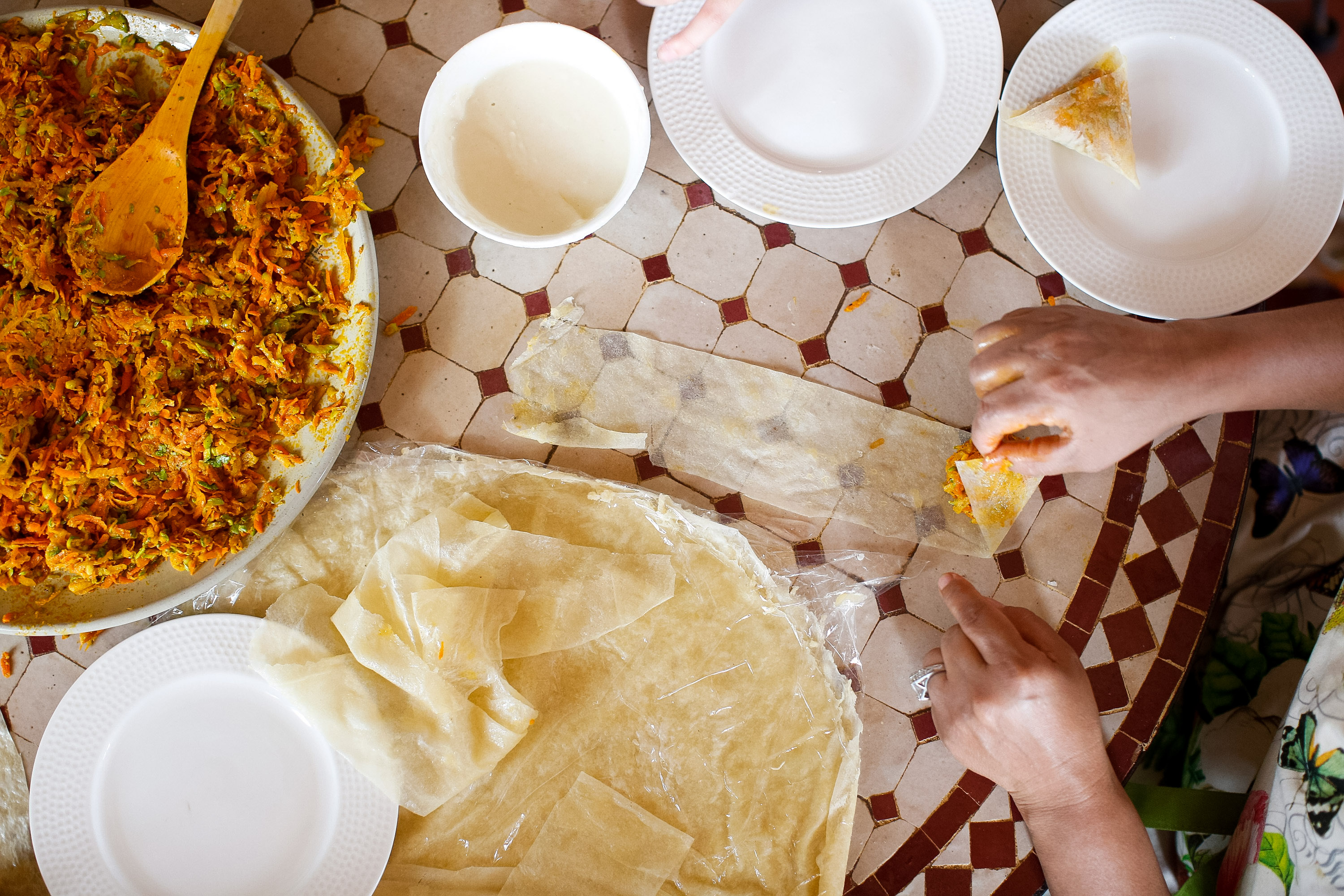 Hands fold thin dough sheets beside a bowl of spiced filling on a tiled table