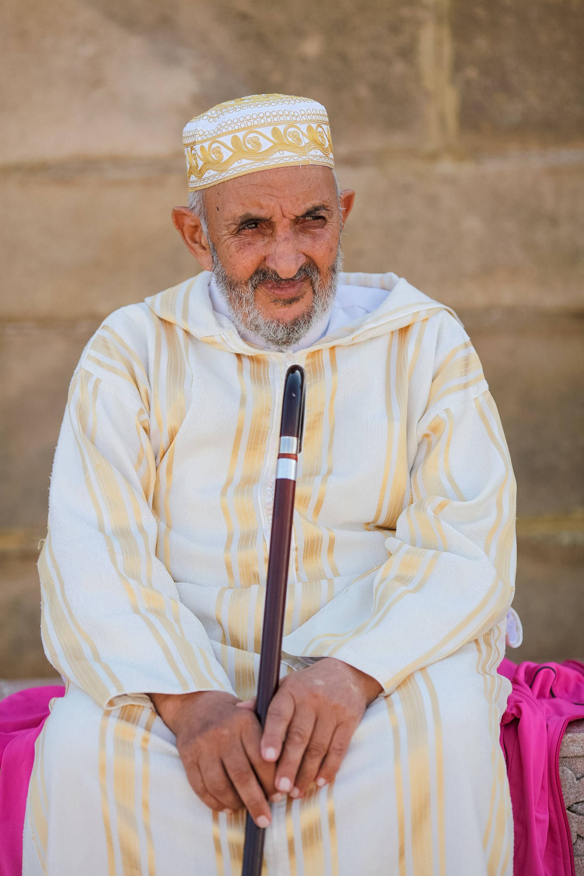 An elderly man in a traditional embroidered cap and robe sits holding a wooden cane