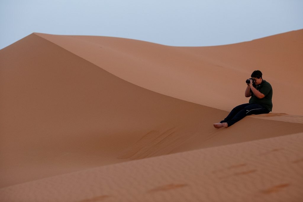 Man sitting on a large sand dune taking photographs.