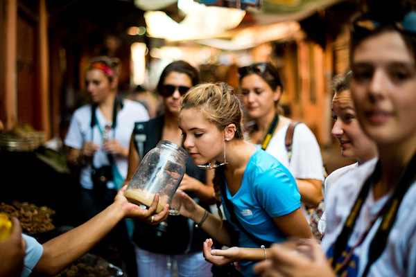Teen travelers lean in to smell a jar held out by a vendor in a crowded market lane in Morocco
