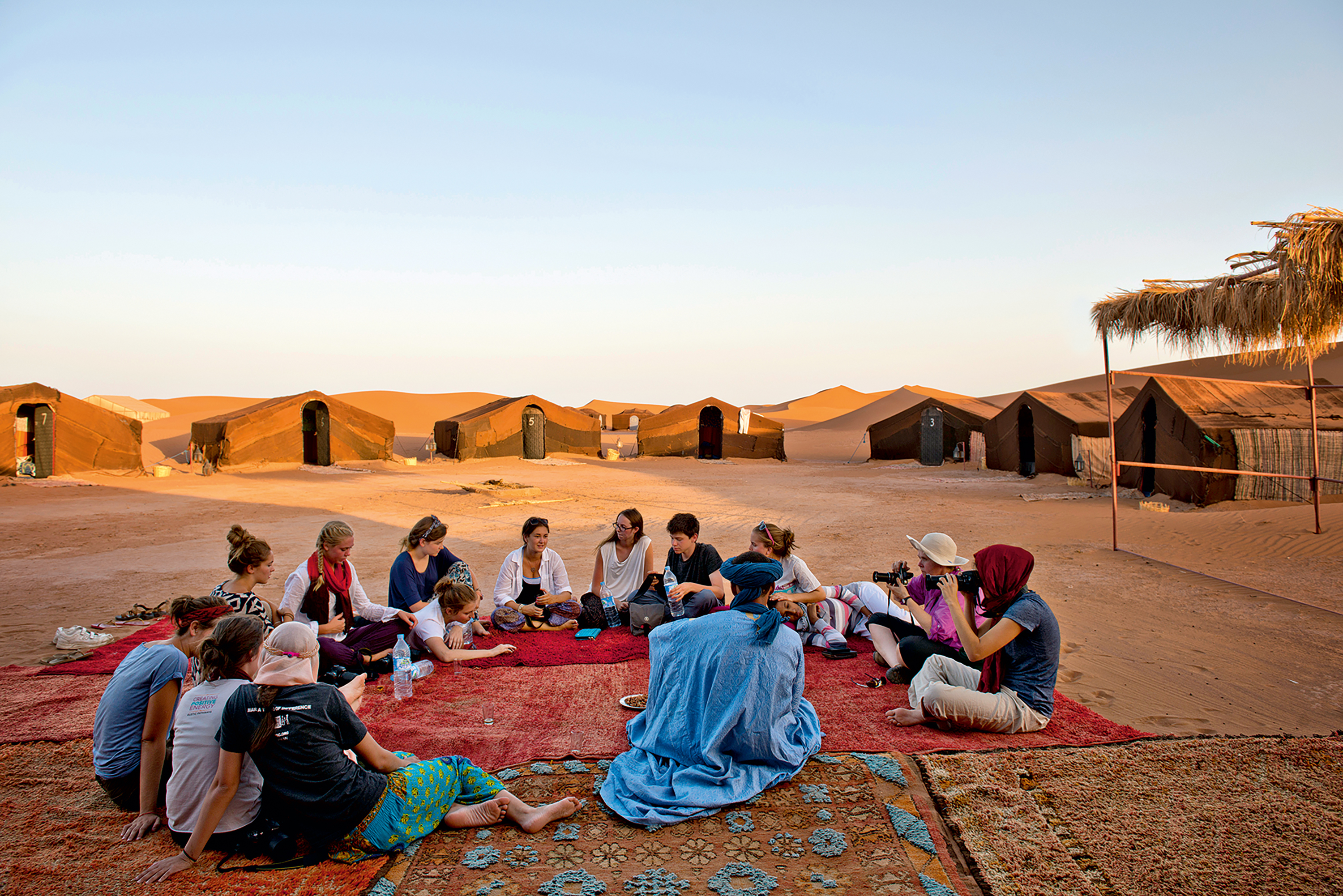 About twenty students sit in a wide circle on patterned rugs spread over the sand, with a line of low dark tented rooms behind them and an orange dune rising beyond.