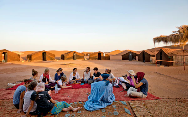 About twenty students sit in a wide circle on patterned rugs spread over the sand, with a line of low dark tented rooms behind them and an orange dune rising beyond.