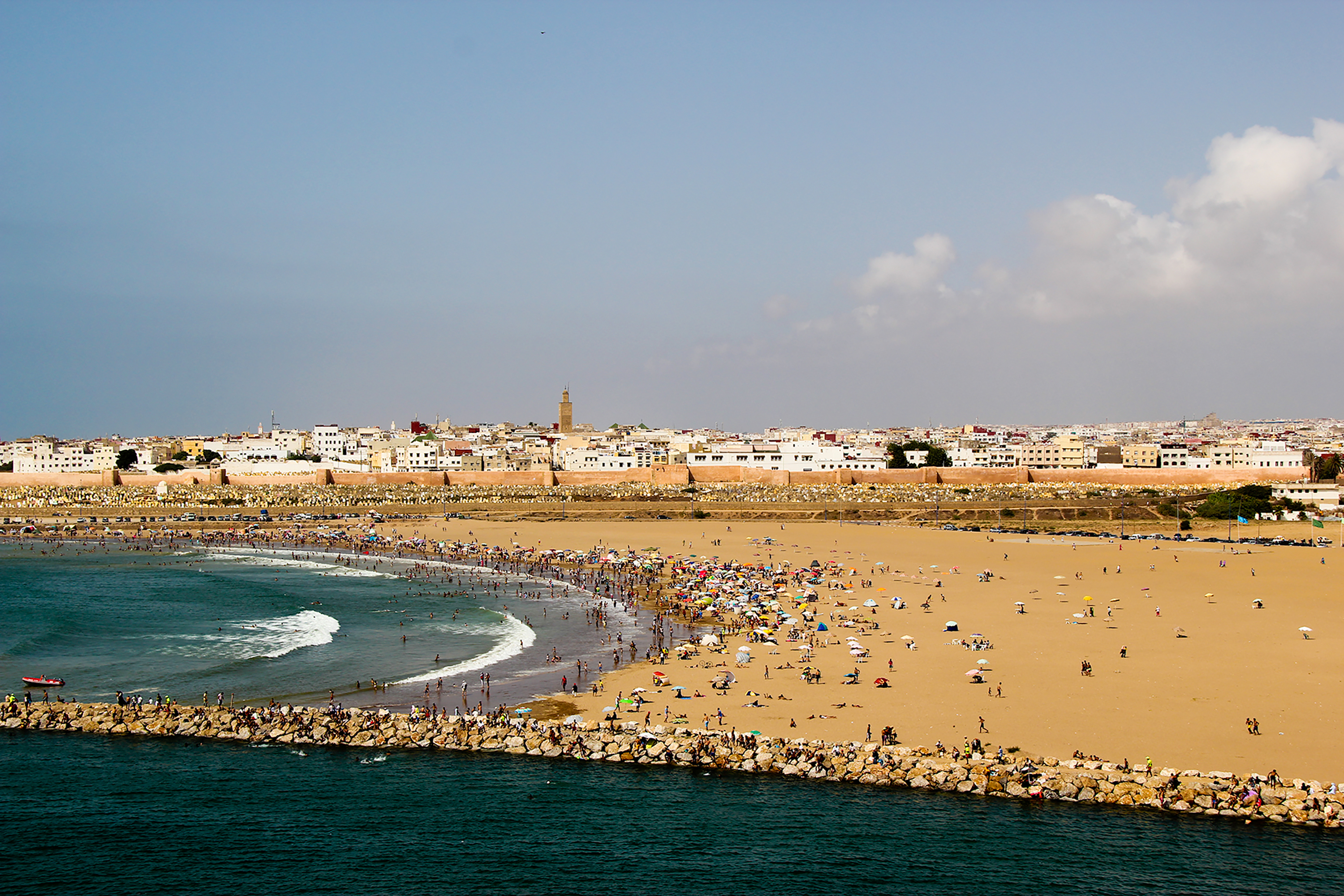 Students walk a crowded beach that curves along a coastal city with whitewashed buildings and a minaret in the distance