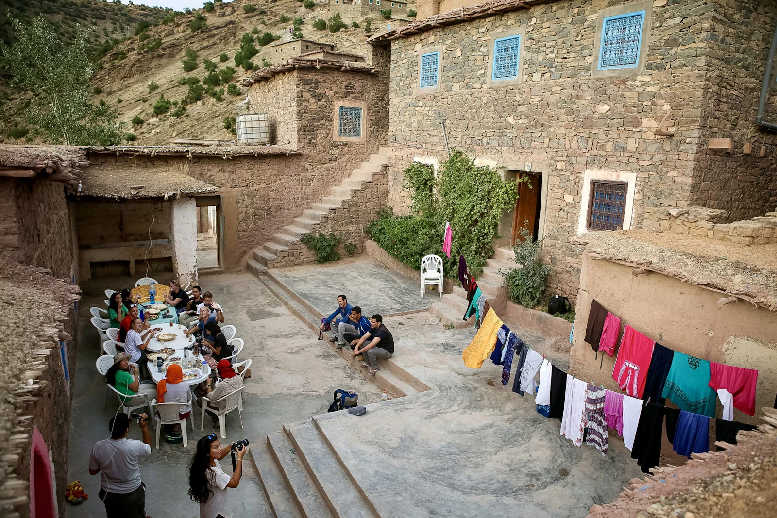 Students and village families share a meal at long tables in the courtyard of a stone house on a hillside in Morocco