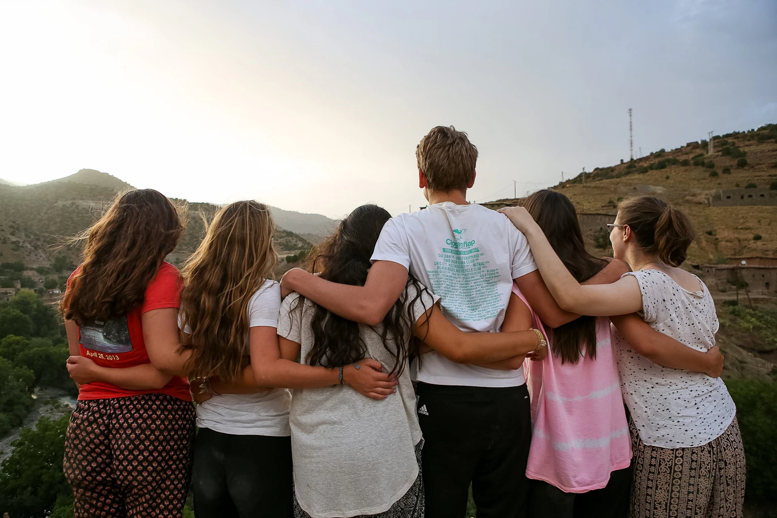 A group of students stands arm in arm with their backs to the camera, looking out over green hills at dusk in Morocco