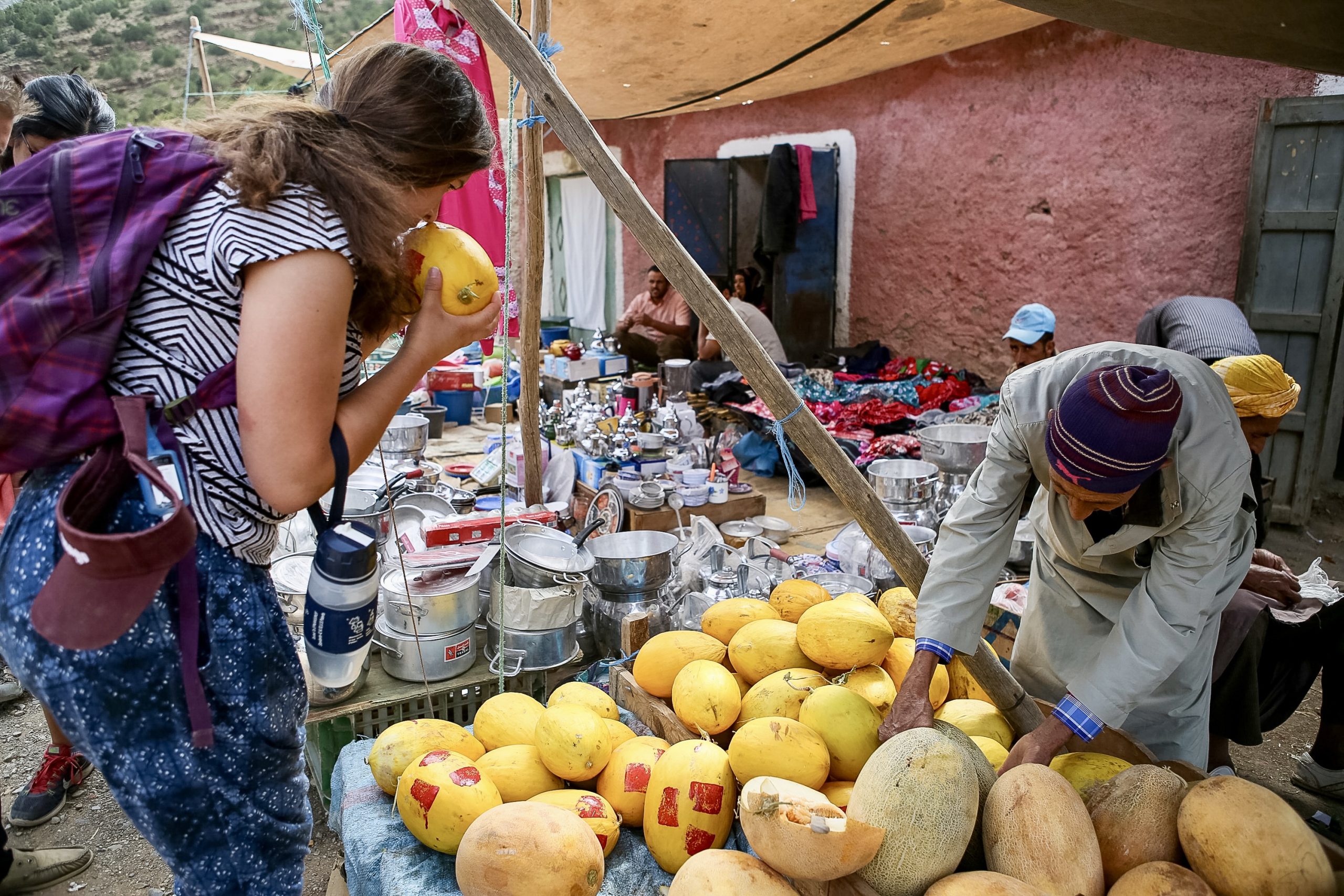 Student smelling fresh melon at an outdoor market stall in rural Morocco, with local vendors selling produce and goods.