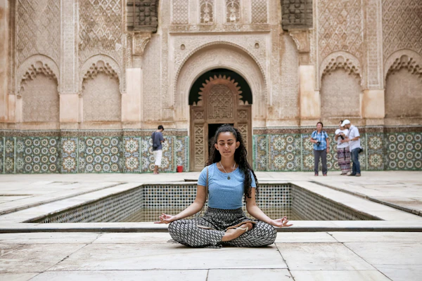 A student sits cross-legged beside a long courtyard pool framed by carved plaster and tiled walls in Morocco
