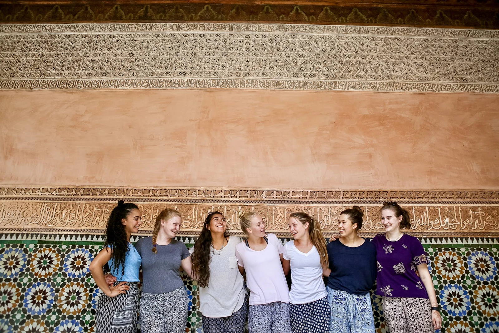 Students laugh together in front of a wall of colorful geometric tilework and carved plaster in Morocco