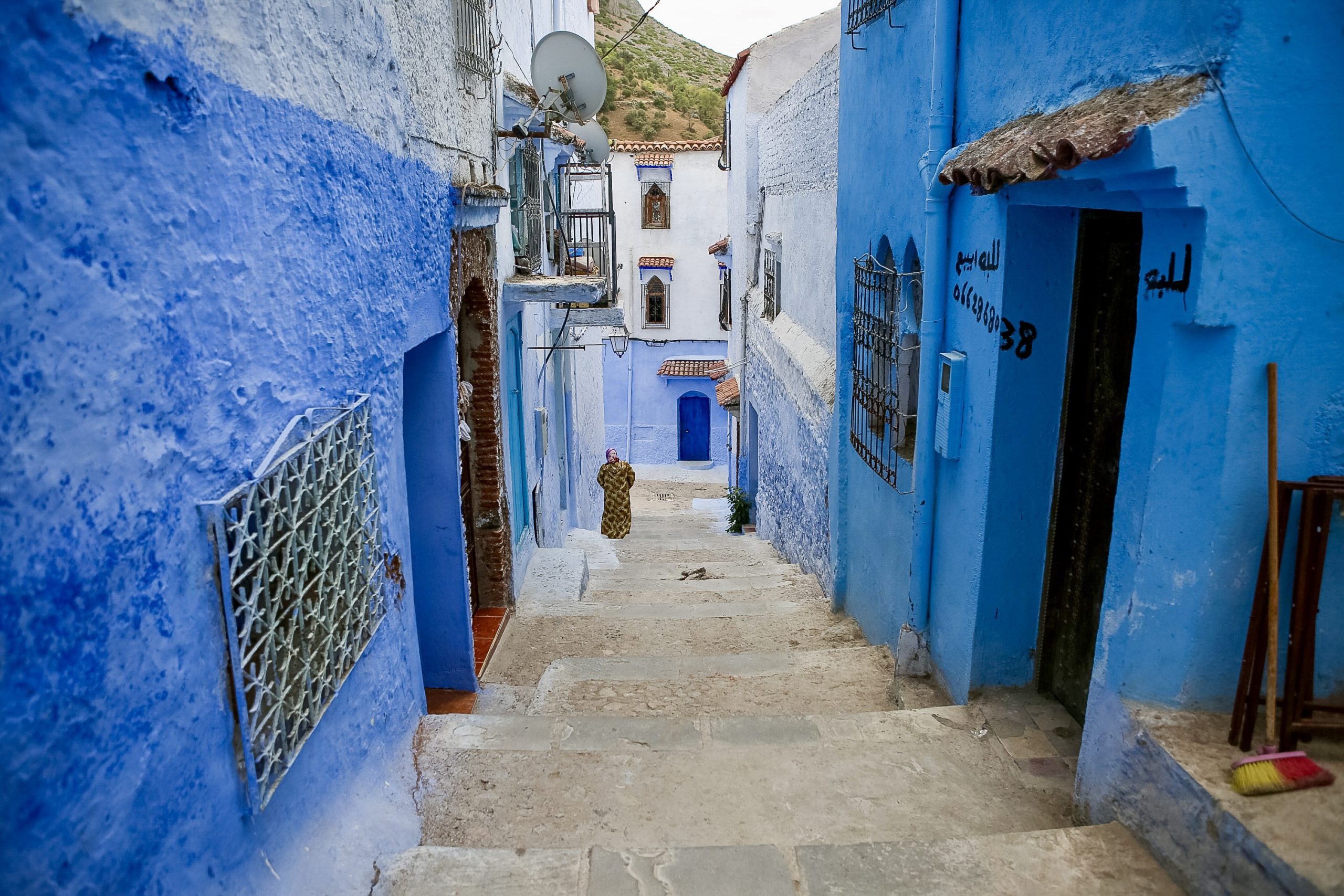 Narrow stone alleyway in Chefchaouen, Morocco, with blue-painted walls and a local resident walking uphill.
