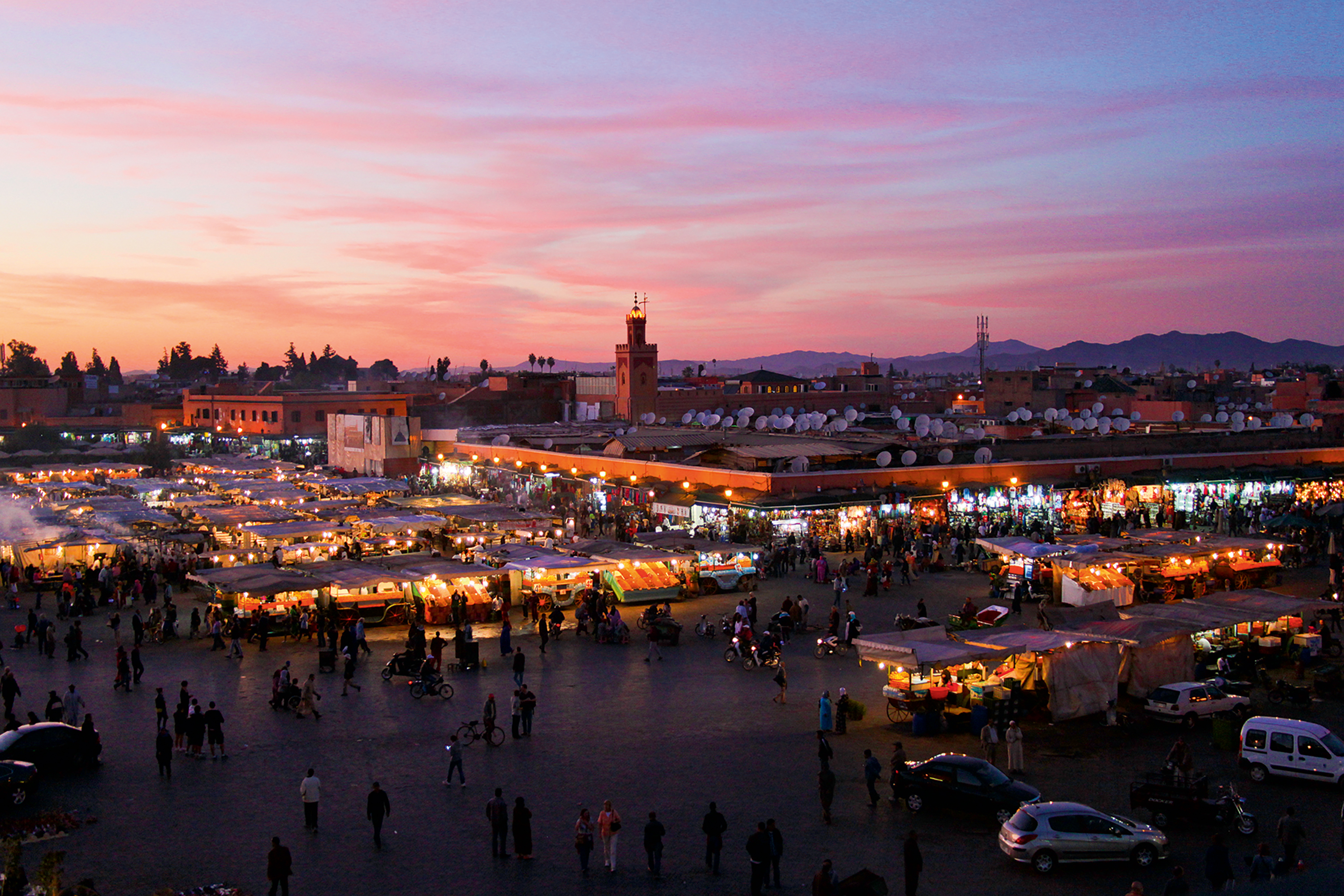 Student travelers pass through Marrakech's Jemaa el-Fnaa market as food stalls glow under a pink sunset sky