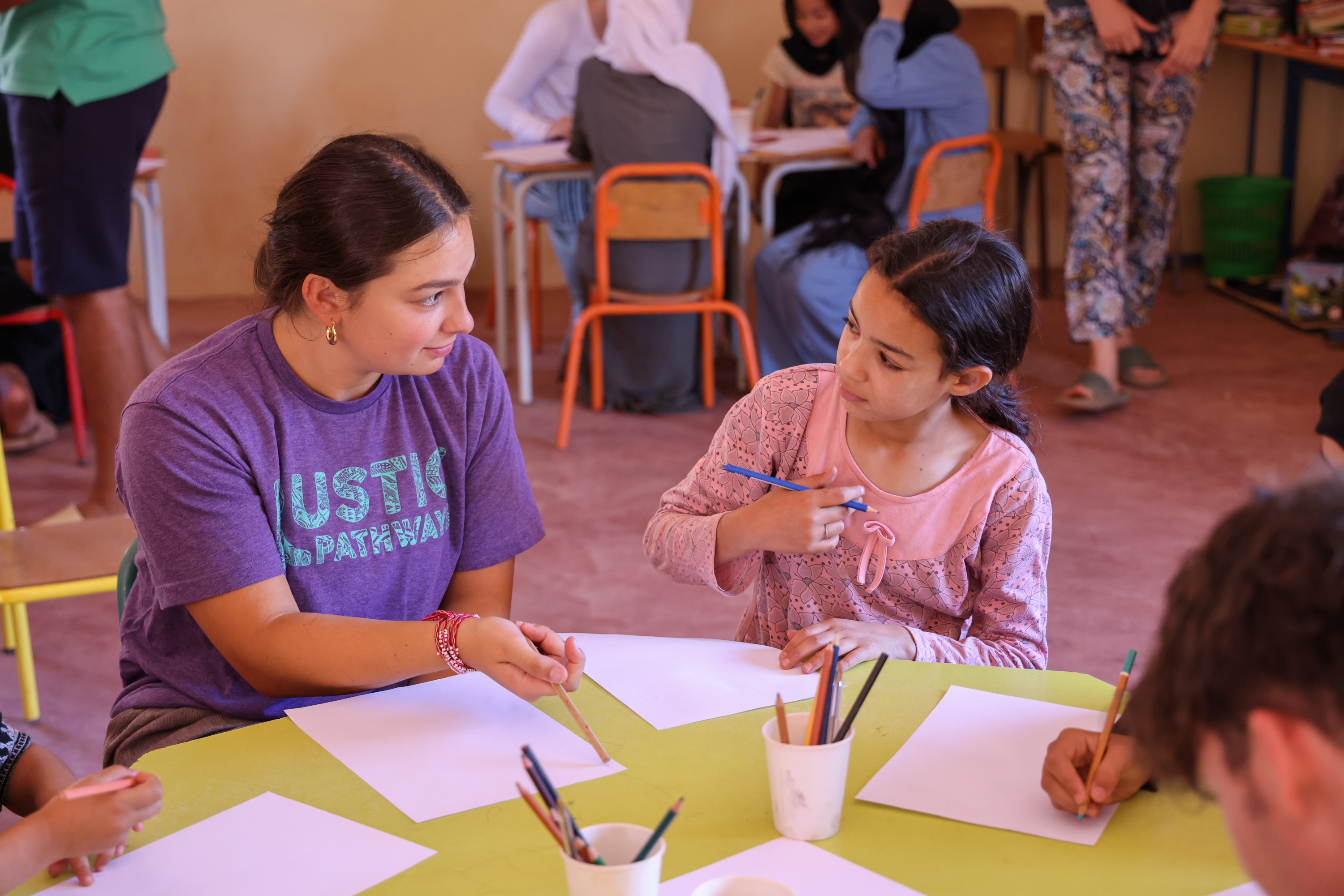 A student in a purple Rustic Pathways T-shirt sits at a classroom table beside a young girl holding a blue colored pencil, with drawing paper and pencil cups between them.